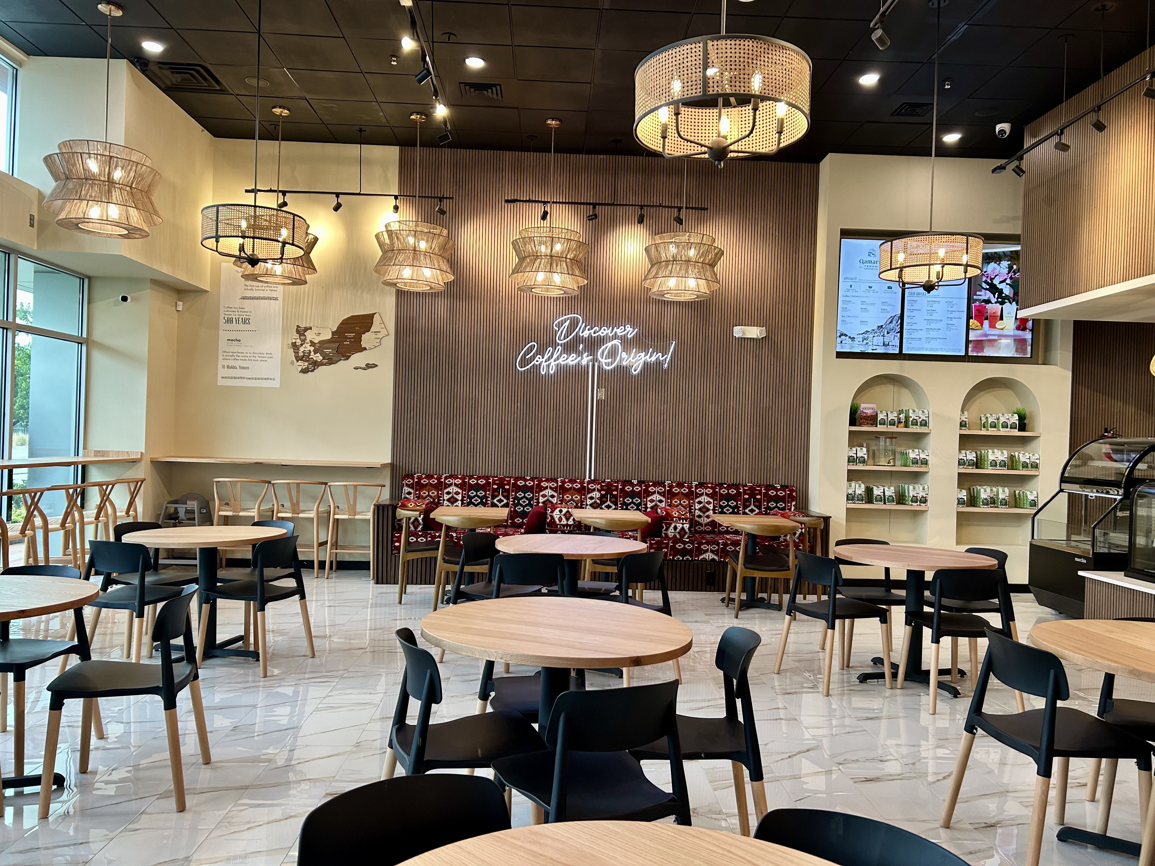 Bright coffee shop interior with round wooden tables, black chairs, colorful bench seating, hanging lattice light fixtures, and a neon sign reading "Discover Coffee's Origin" on a striped wooden wall.