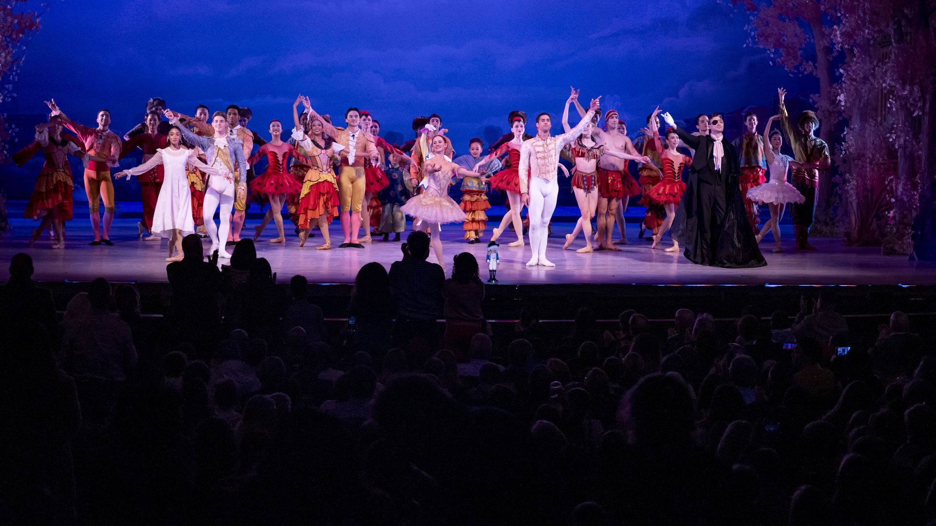 Wide shot of a ballet finale: many dancers in bright period costumes on a blue-lit stage, center couple in white, surrounded by red, yellow, and pink outfits; audience silhouettes foreground.
