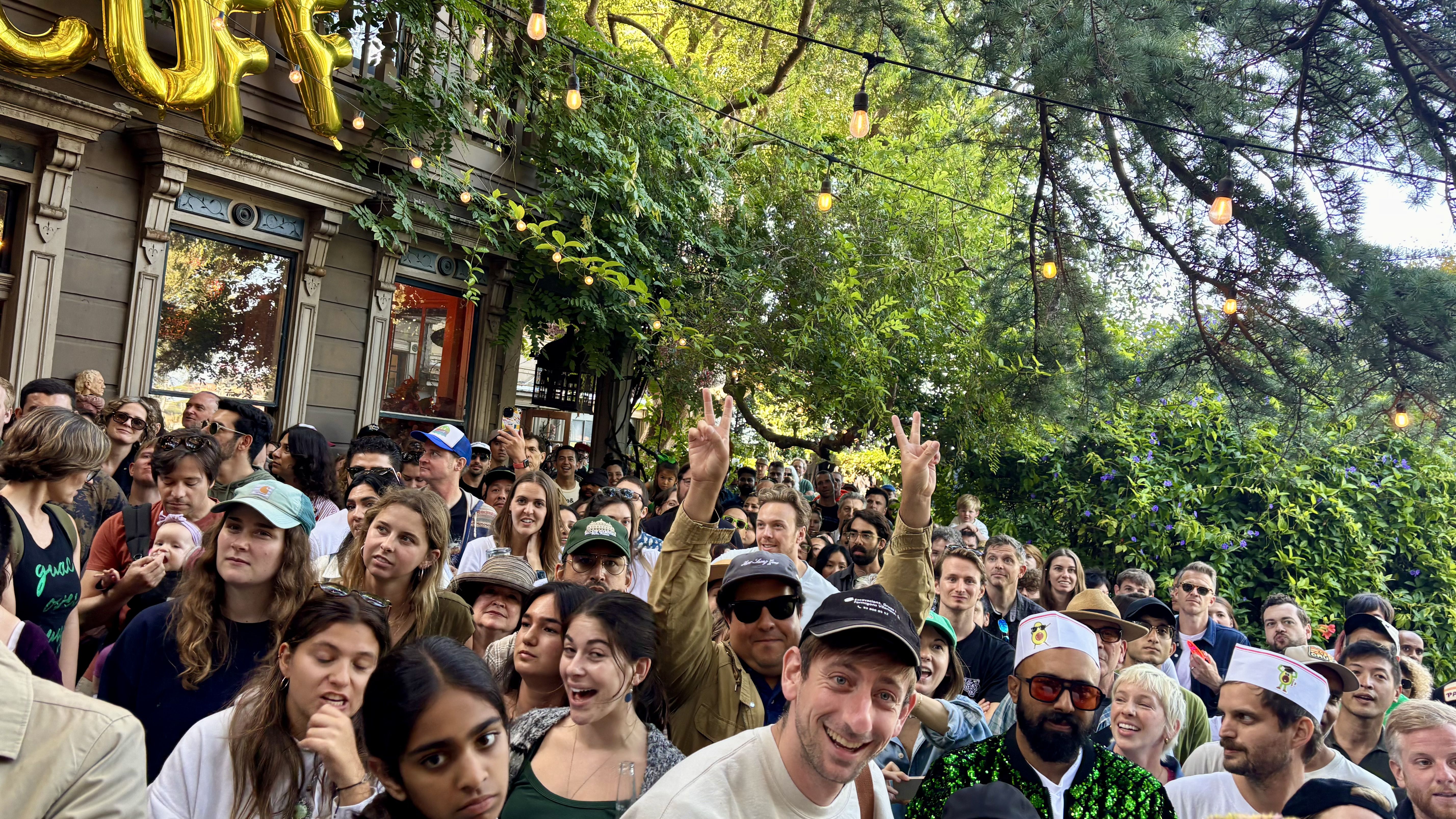 Crowd of diverse people gathered outdoors near a building with gold balloons and string lights, surrounded by trees and greenery, some wearing hats and smiling, lively daytime event.