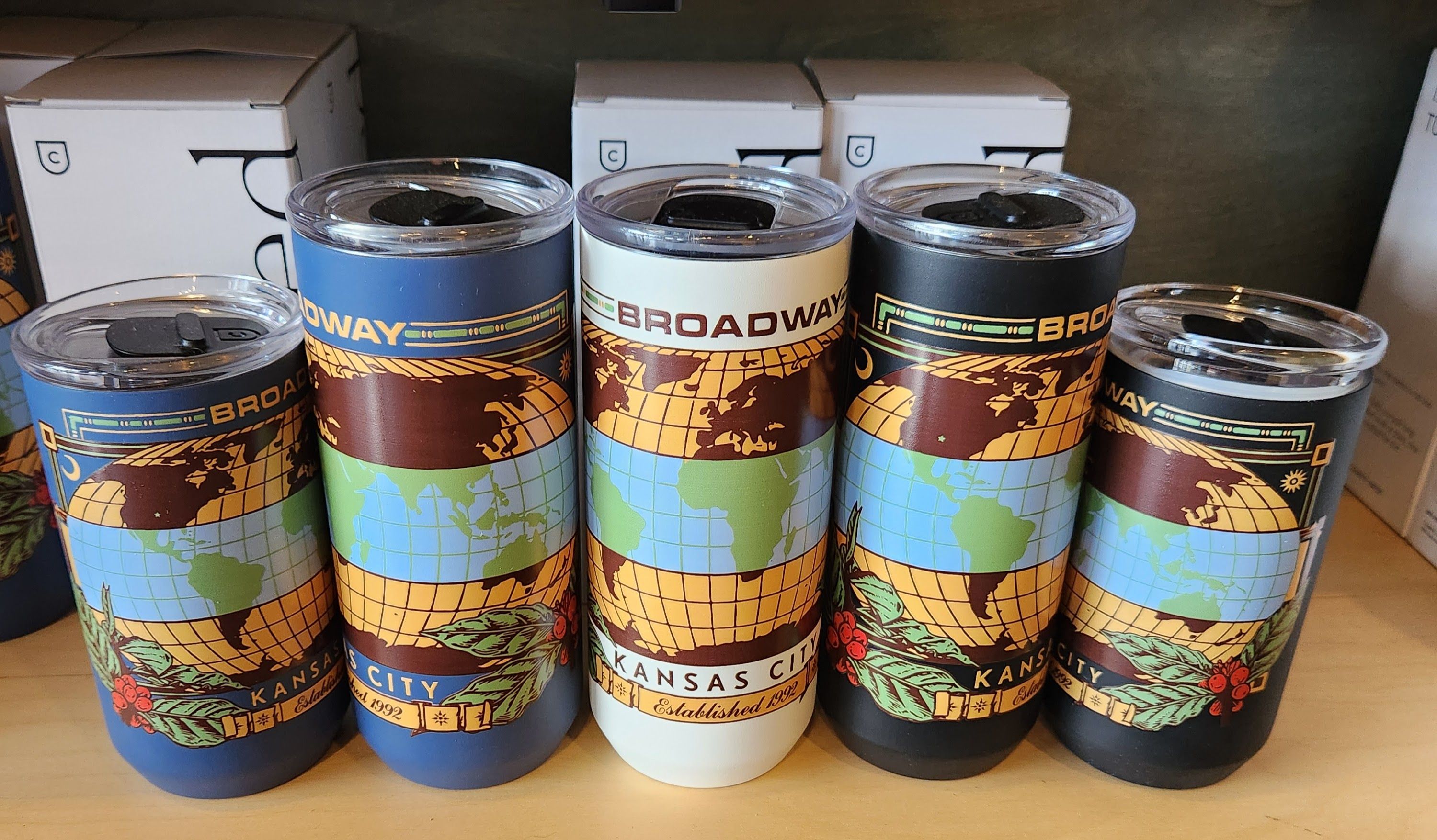 Five cylindrical travel mugs with world map graphics and the words 'Broadway' and 'Kansas City' stand side by side on a shelf; white boxes with a C logo sit behind them.