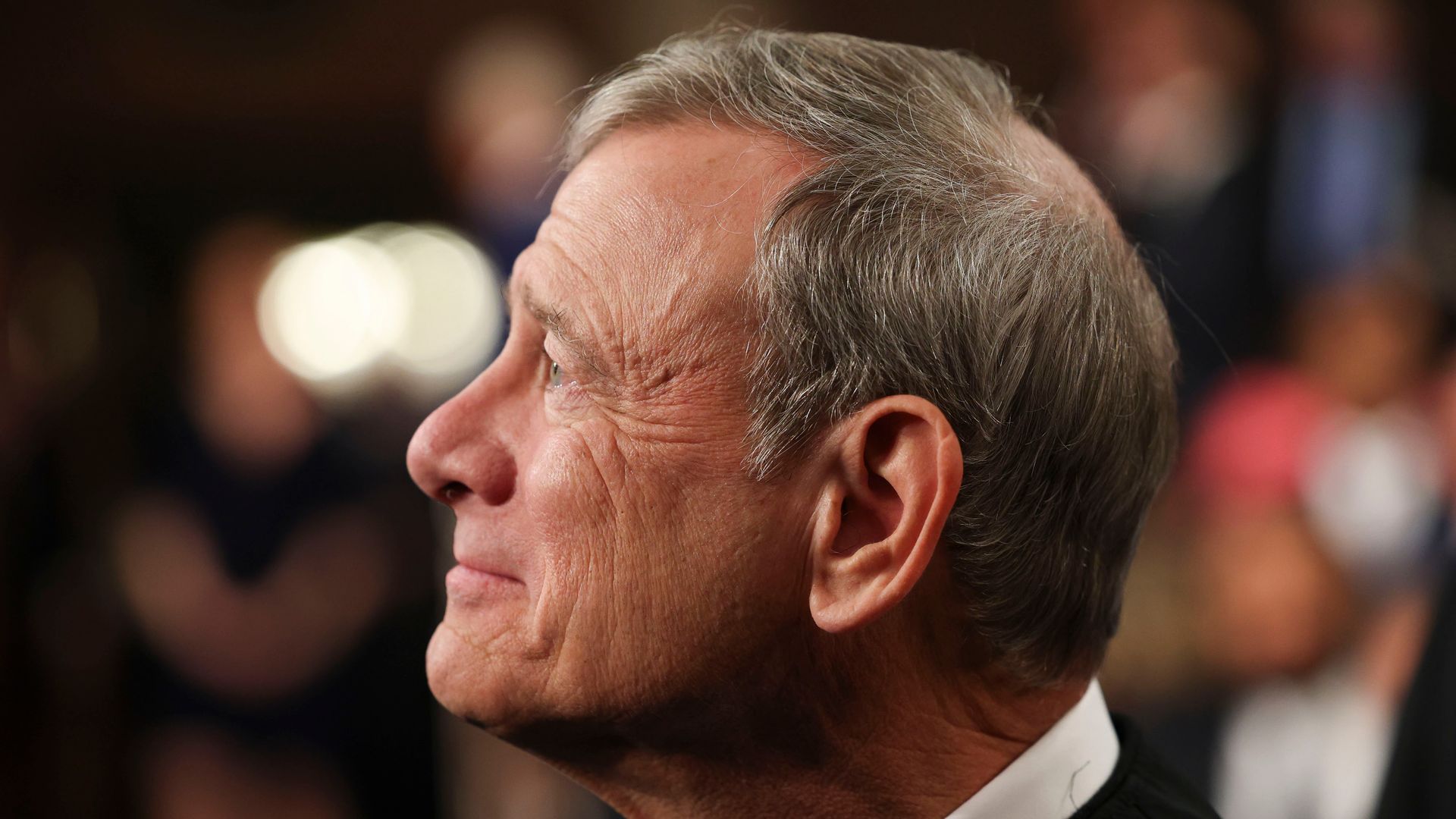Side profile of a gray-haired man in a black suit and white shirt, looking to the left; out-of-focus crowd and soft lights blur the background.