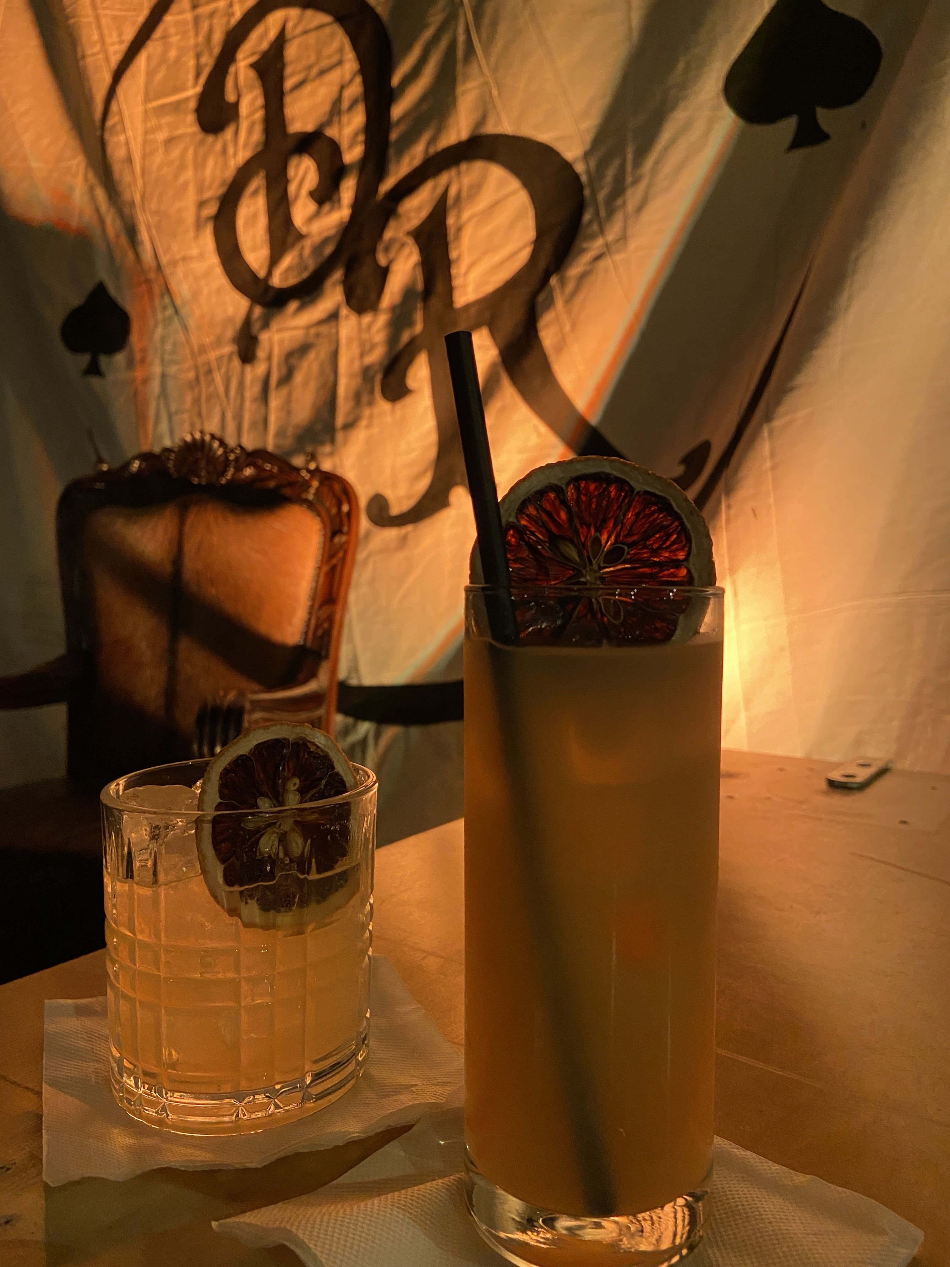 Two cocktails with dried citrus garnish on a wooden table, warm orange lighting, ornate chair in background, and draped fabric with black card suit designs and large script letters.