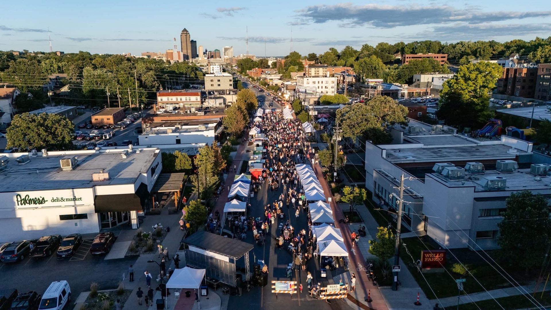 An aerial view of a city street festival: white tents line a closed road with crowds, shops along the sides, trees, and a distant skyline under a blue sky.