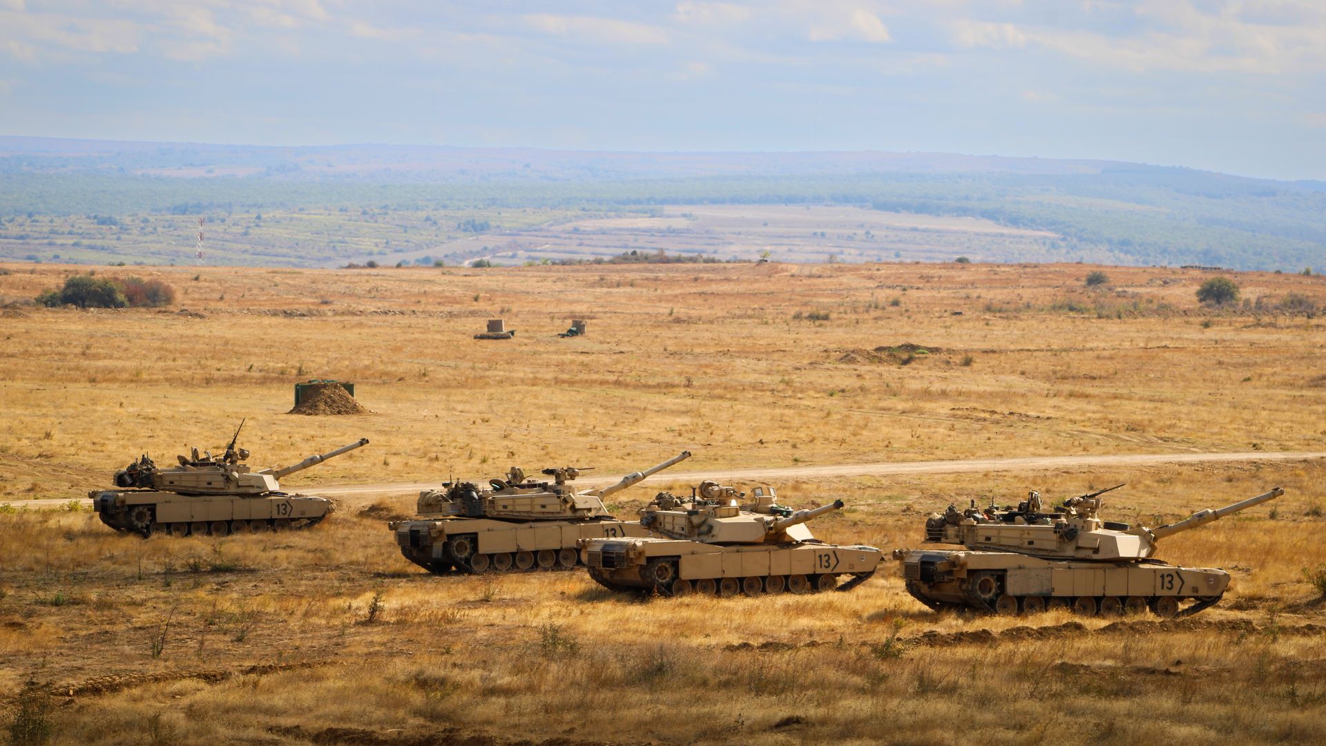 Four tan military tanks positioned in a dry, open field with rolling hills and a cloudy sky in the background.