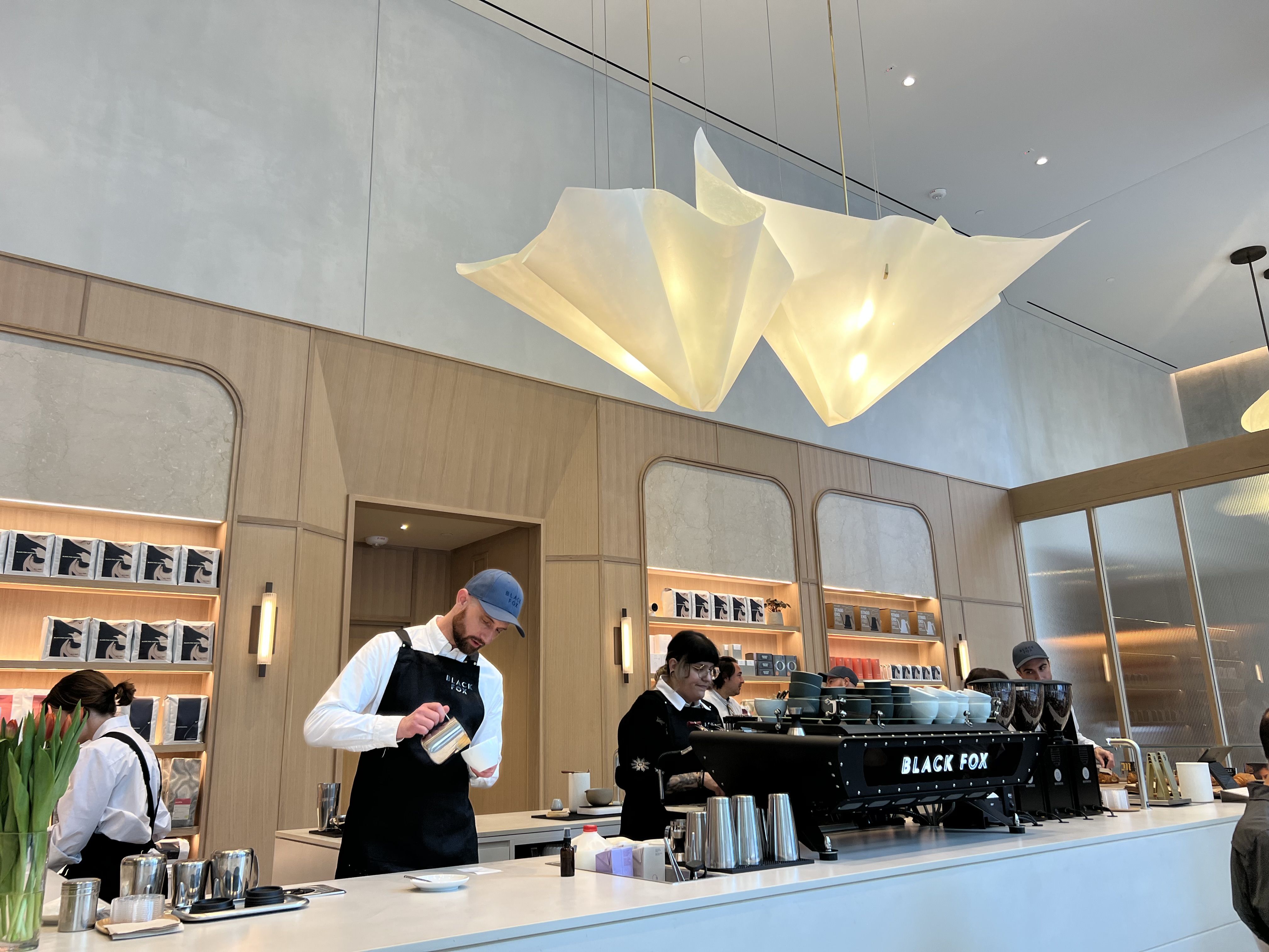 Interior of a bright cafe featuring a large white origami-style chandelier above the counter; baristas in aprons work at a Black Fox espresso machine, with wood paneling and shelves behind.