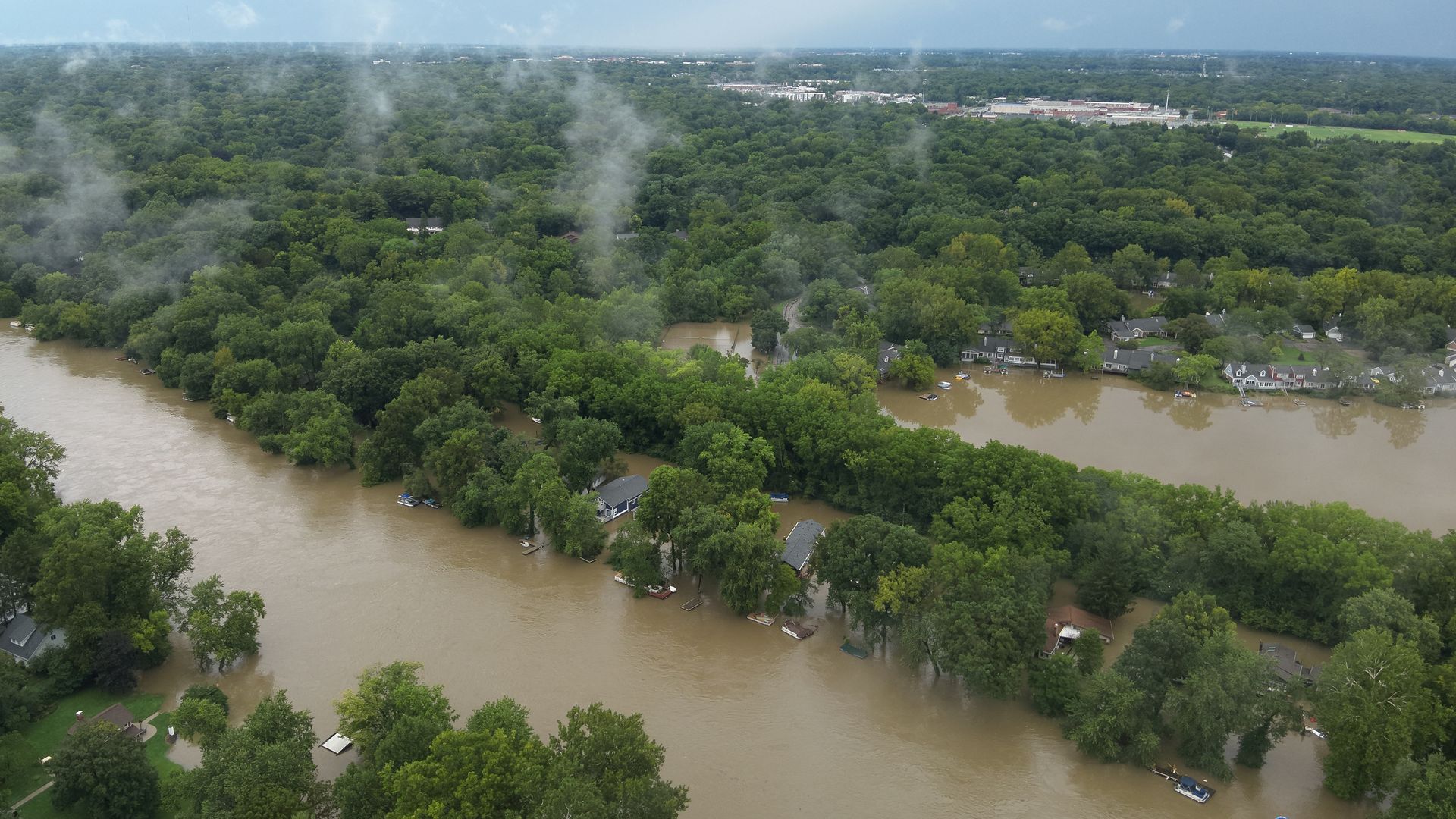 An aerial panorama of a muddy river flooding a dense green forest, with houses along the banks and a distant town on the horizon; wispy smoke rises from the trees under a blue-gray sky.