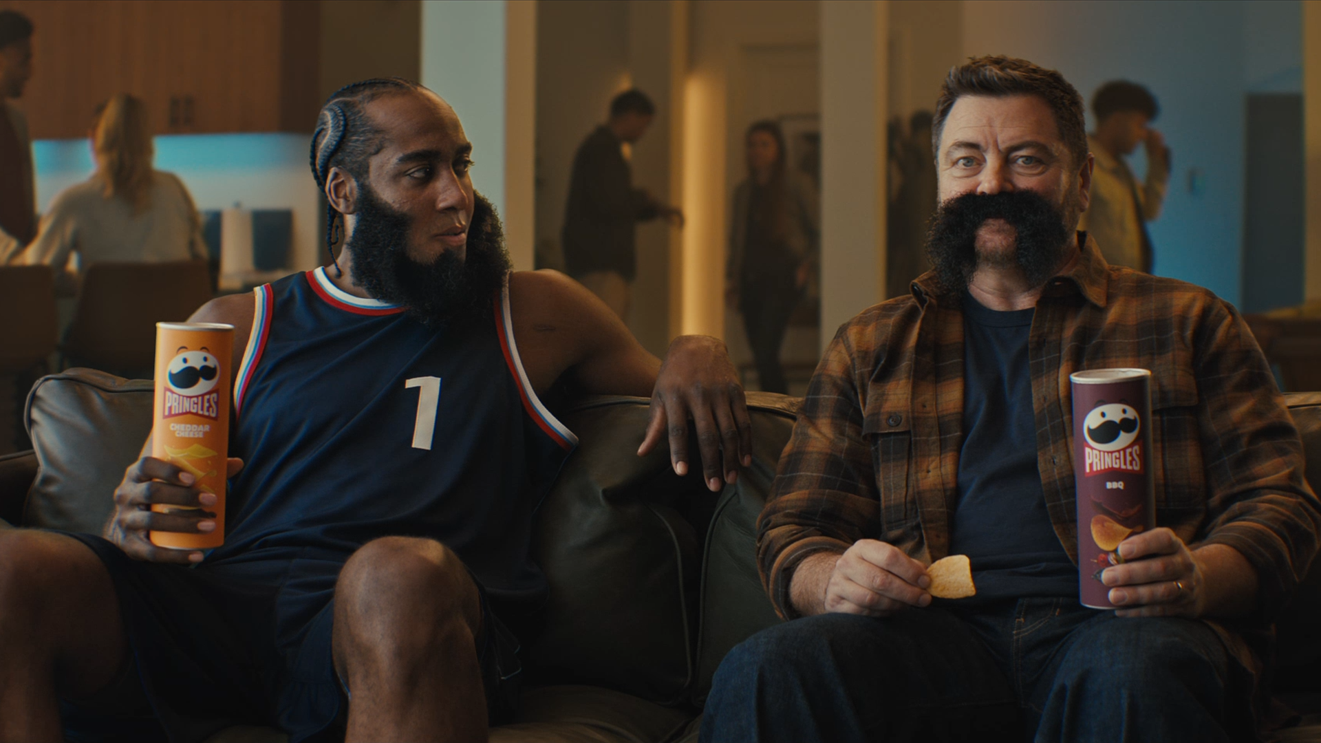 A photo shows James Harden holding a Pringles can sitting on a couch, looking next to him at Nick Offerman, who is wearing his mustache.