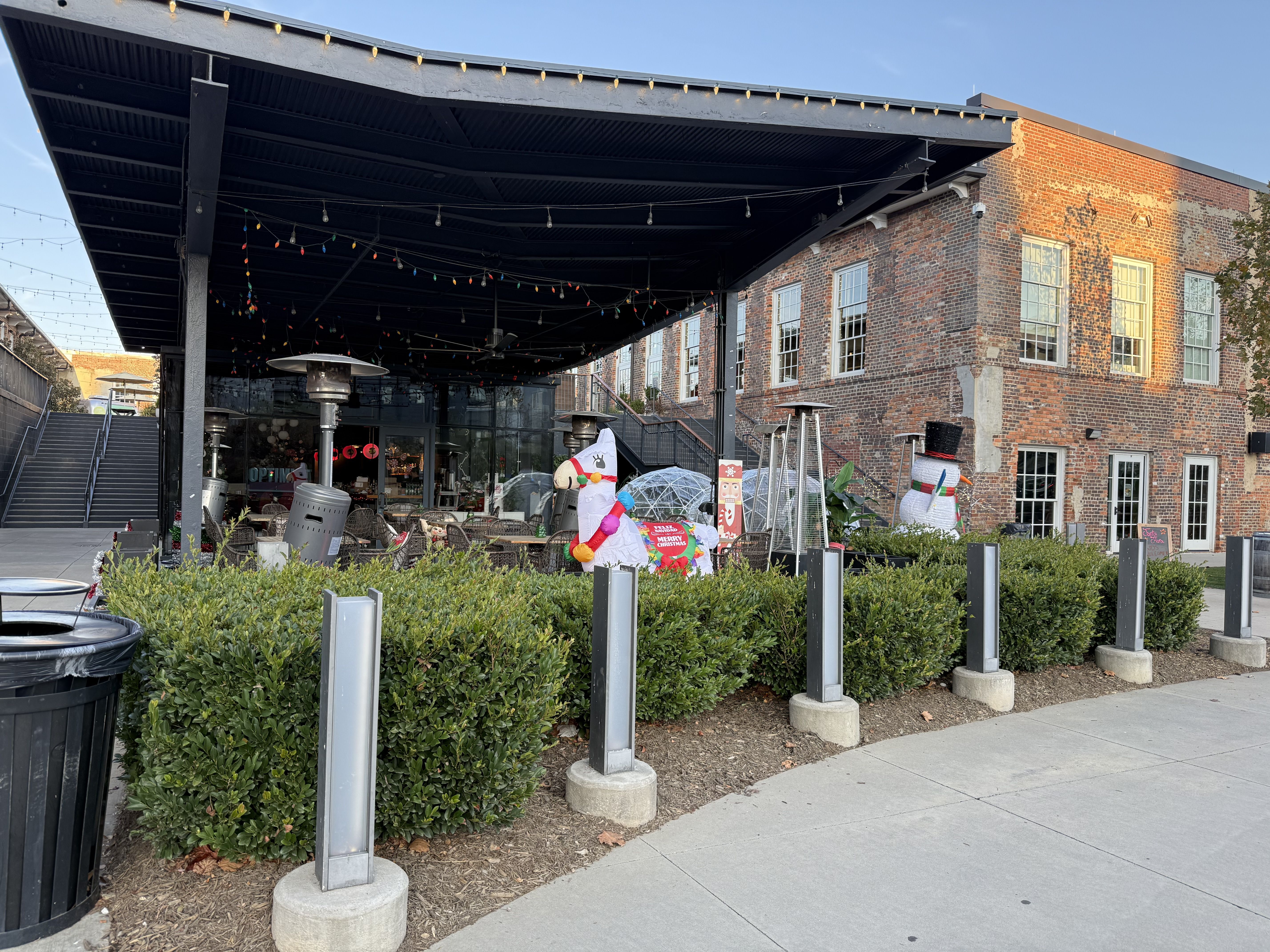 Outdoor patio area with black roof, holiday decorations including inflatable llama and snowman, string lights, green hedges, brick building, and clear winter sky.