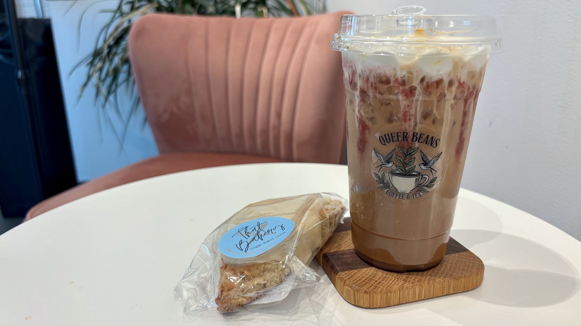 Iced coffee in a clear cup with a plastic lid, cream swirls and fruit pieces visible; logo reads "QUEER BEANS COFFEE & TEA" on the side, on a white table with a pink chair and plant behind.