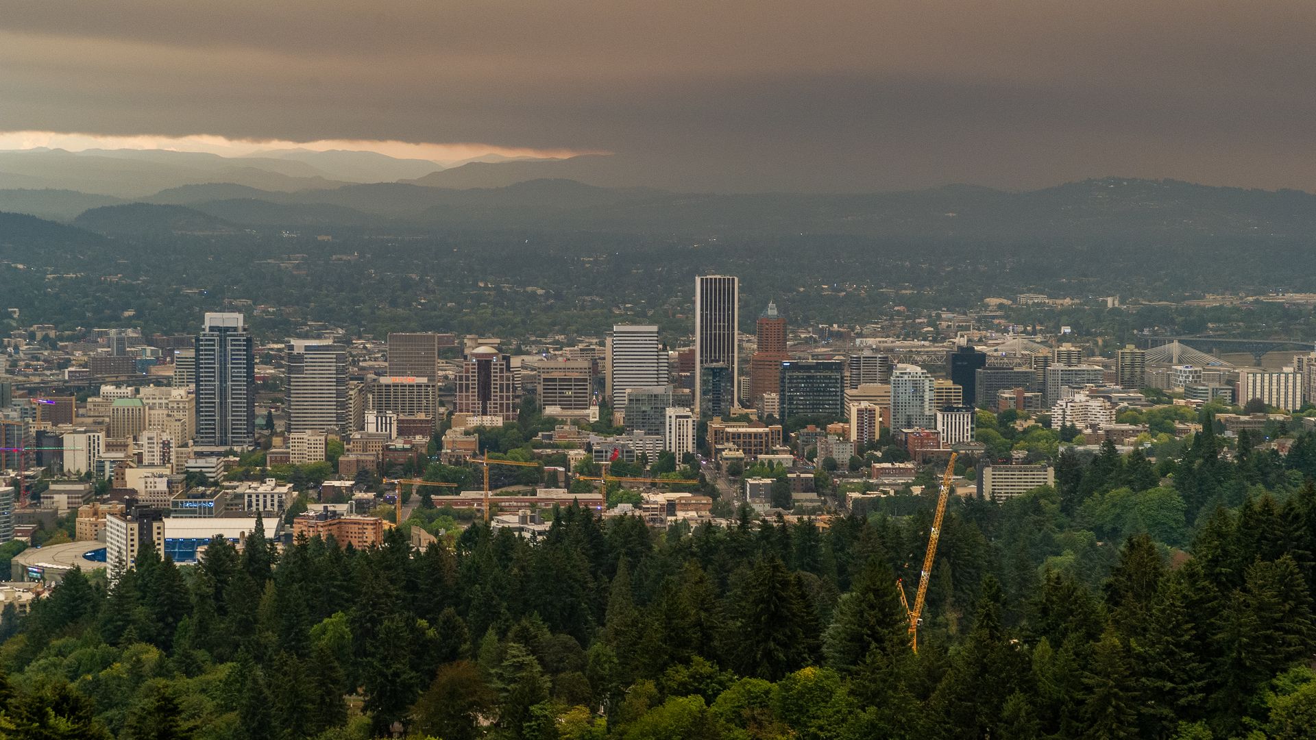 View of the Portland skyline with tall buildings behind a dense green forest, under a dramatic brown and gray cloudy sky filled with wildfire smoke, with distant mountains on the horizon.