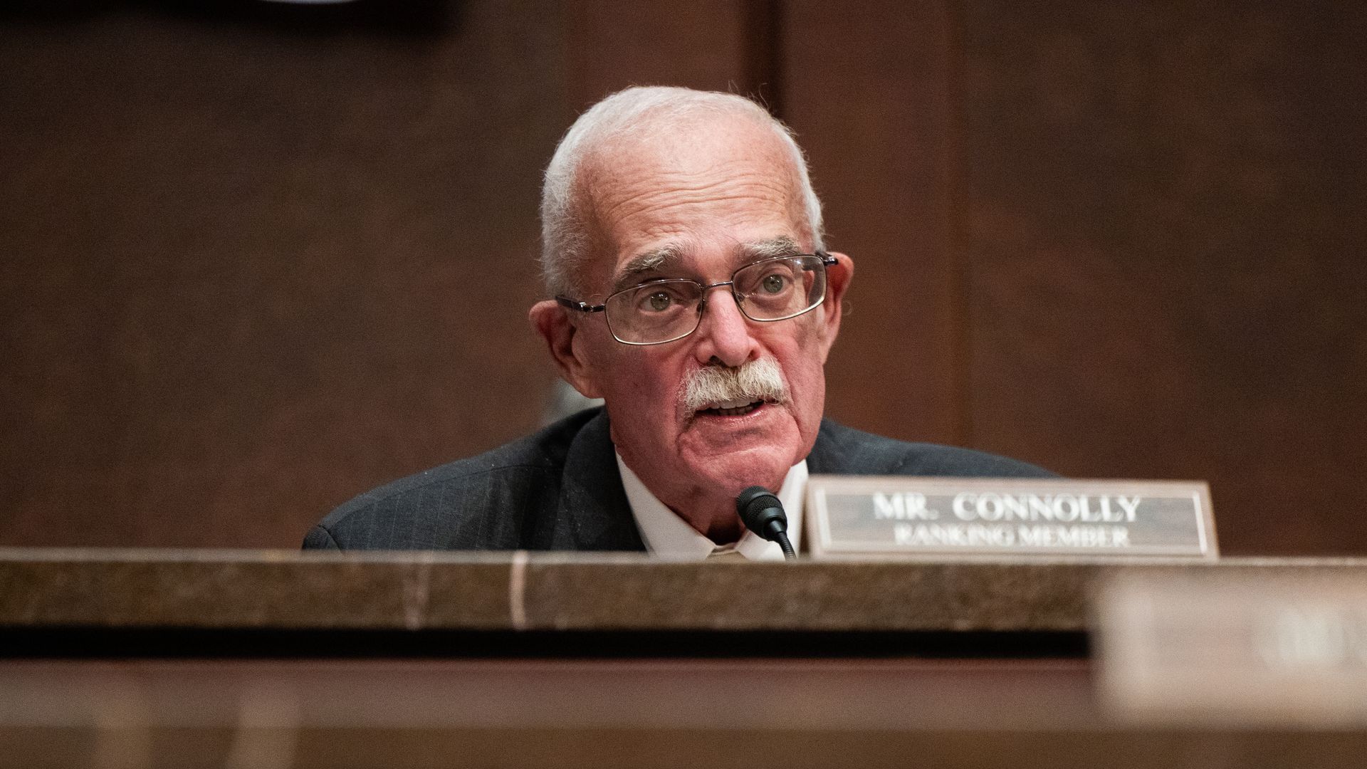Rep. Gerry Connolly, wearing a gray suit and speaking into a microphone while seated at a marble table in front of a brown wooden wall.
