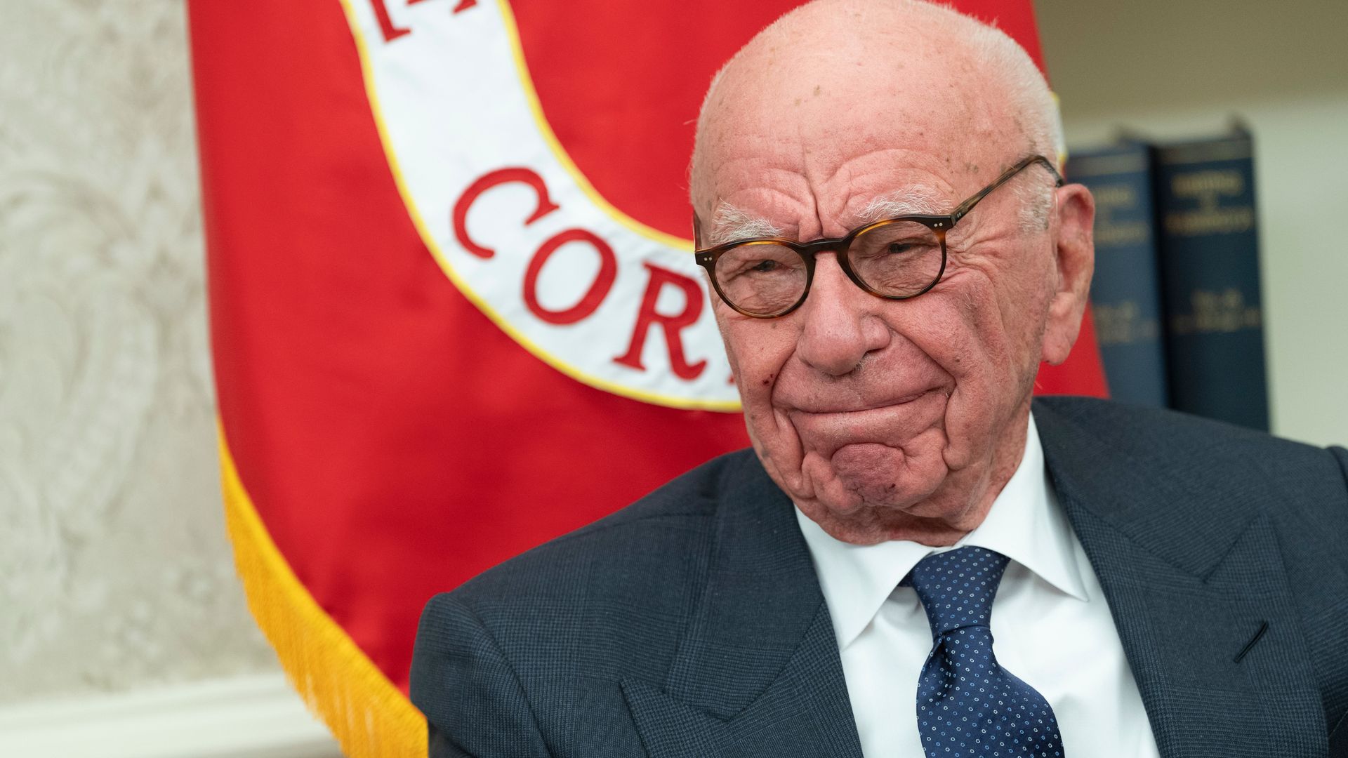 Ruper Murdoch wears a grey suit with a white shirt and a dotted blue tie. He is smiling and wearing glasses.