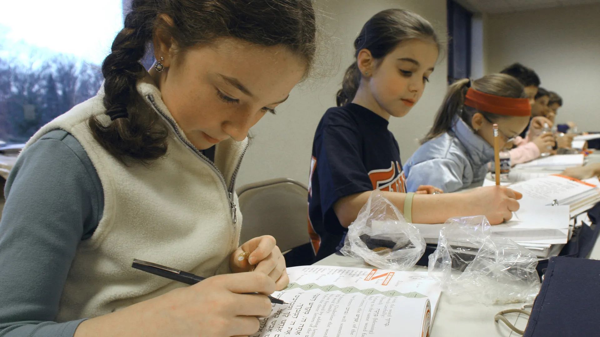 Photo shows girls sitting at a table. They are holding pencils and pens and working on Hebrew notebooks.