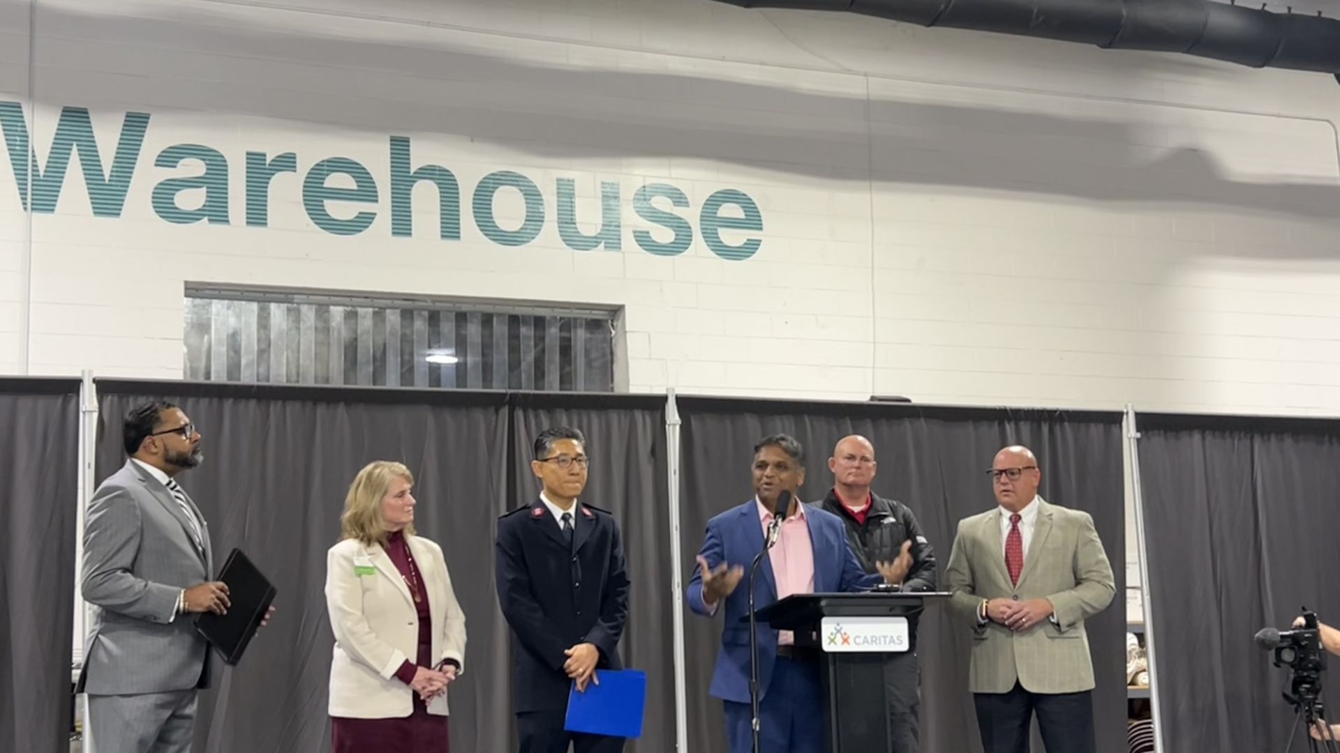 Six people stand in a row inside a warehouse with large teal "Warehouse" sign overhead. One man in a blue suit speaks at a podium labeled "Caritas" while others listen.