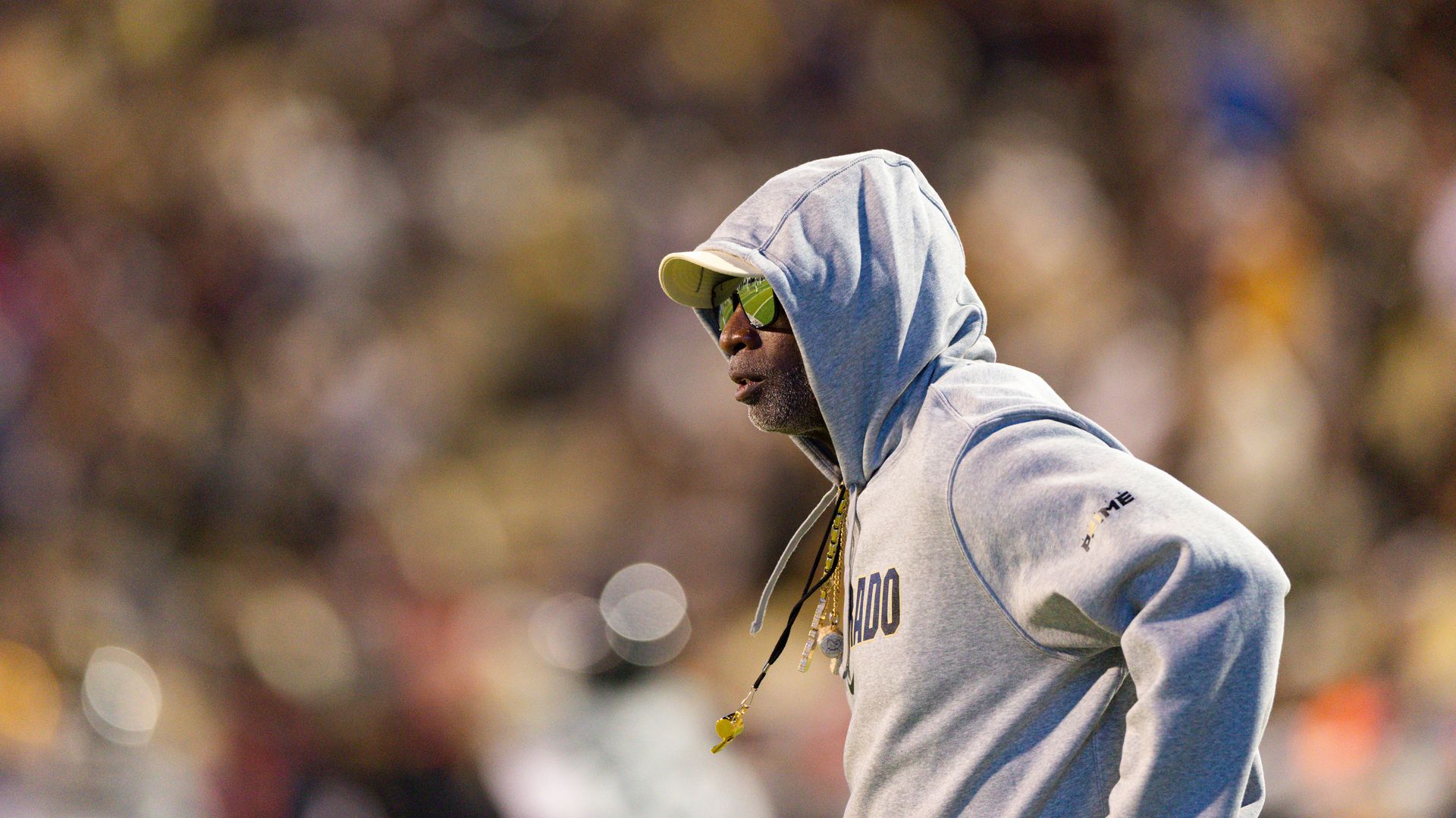 Deion Sanders wearing a light gray hoodie and cap with reflective green sunglasses and a gold chain, standing in a large blurred stadium crowd.