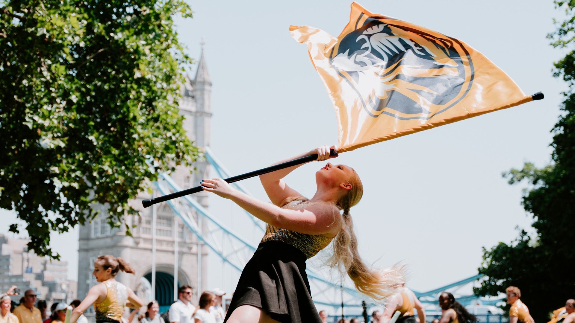 Female performer in a parade waves a large orange flag with a bold black and white emblem. Bright sky, green trees, a distant tower, and a cheering crowd frame the scene.