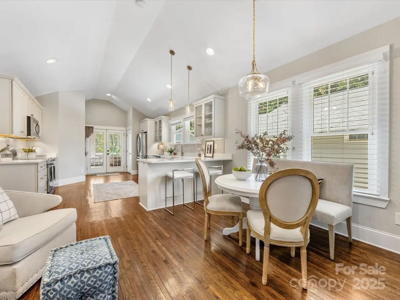 Bright open kitchen and dining area with white cabinets, wood floors, round white dining table, beige chairs, pendant lights, large windows with blinds, and French doors leading outside.