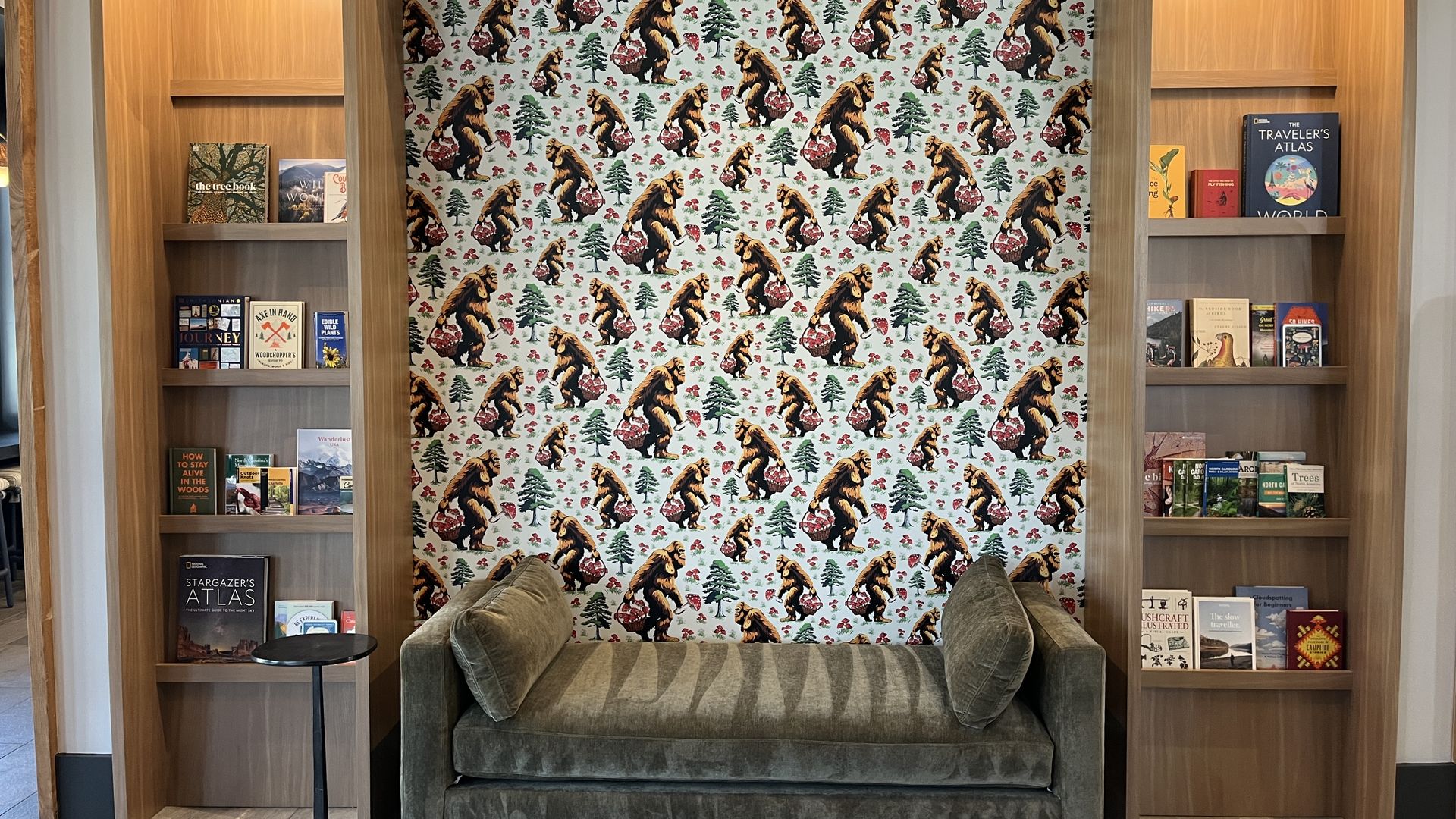 Cozy reading nook with a greenish-gray velvet sofa flanked by tall wooden bookshelves. A wallpapered back wall shows repeating bears and pine trees; books line the shelves.