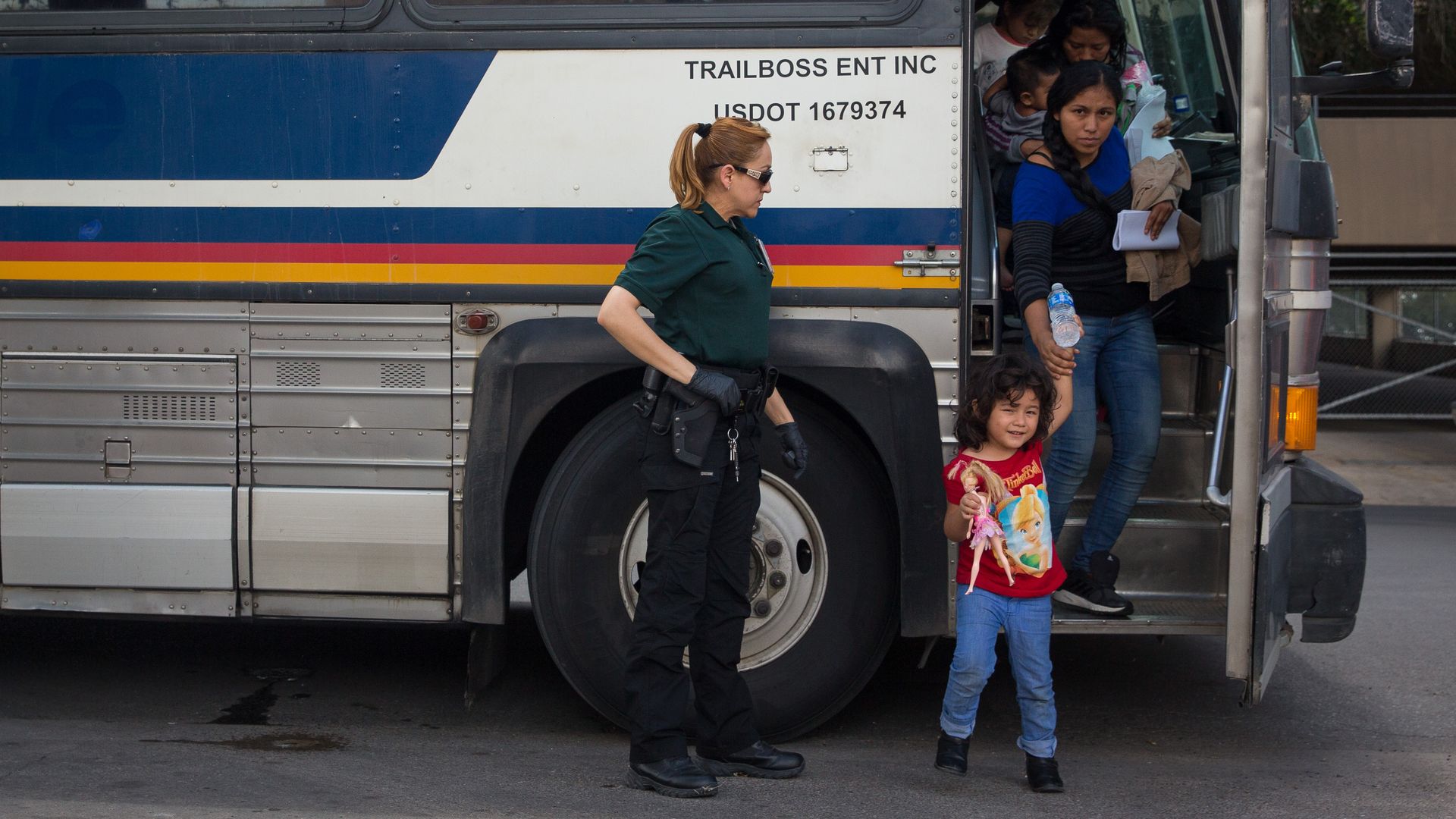 In this image, a young girl steps off a bus with her mother holding her hand while carrying another child. A guard watches.