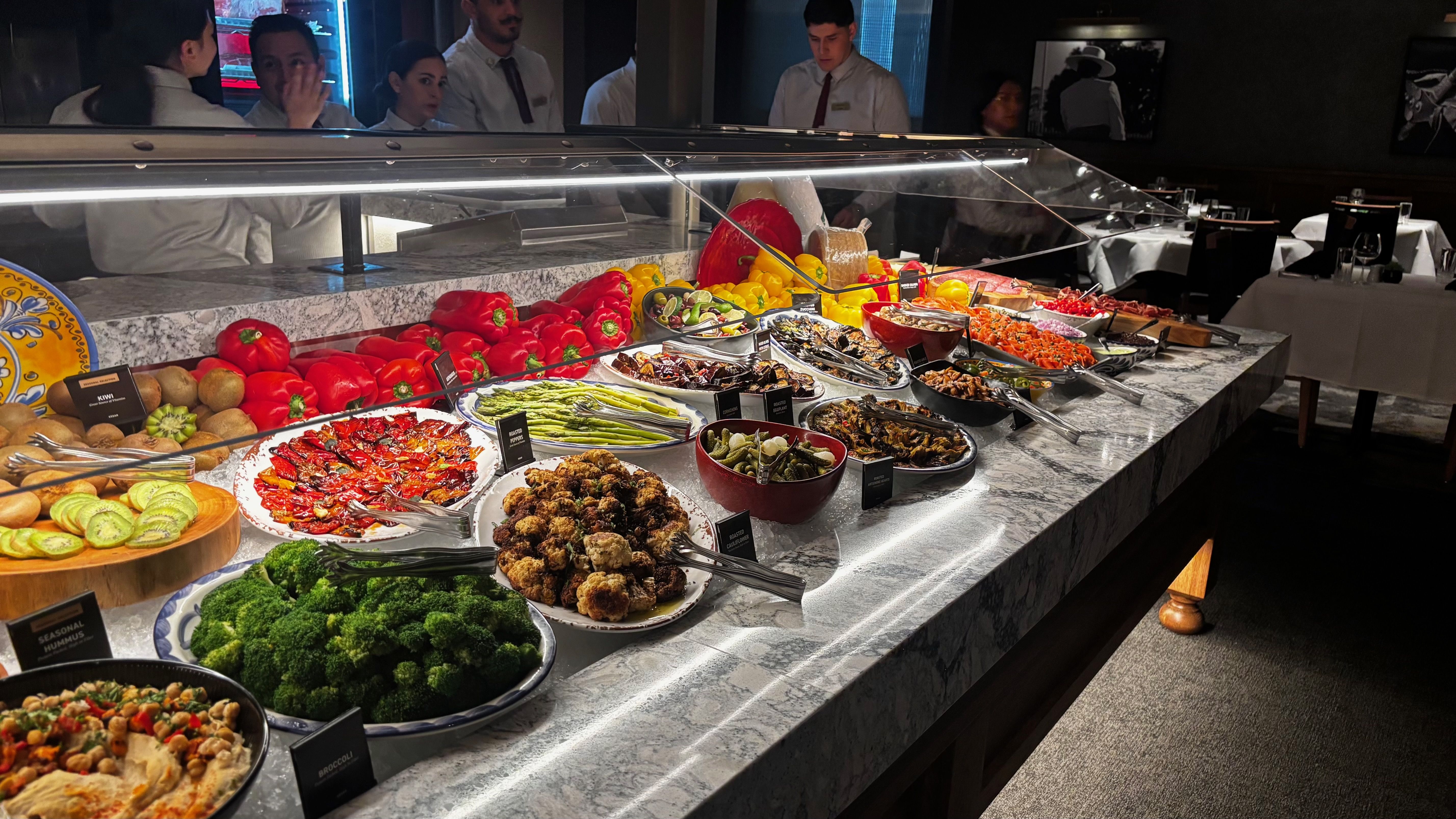 Buffet with fresh vegetables and dishes like broccoli, roasted cauliflower, asparagus, and peppers, displayed on a marble counter with serving utensils and staff in white shirts behind.