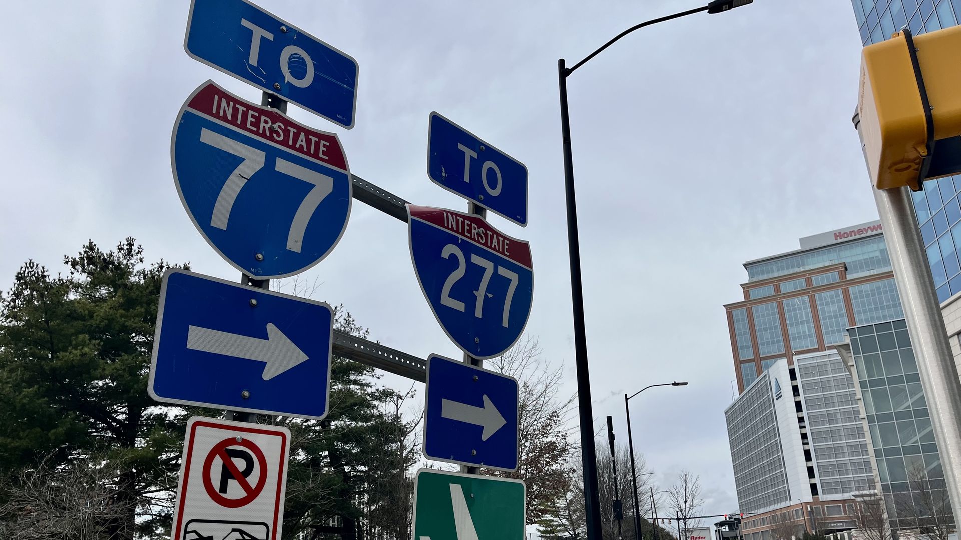 Road signs directing to Interstate 77 and 277 with arrows pointing right, a no parking sign with towing info, and a green airplane symbol sign next to a city street with buildings.