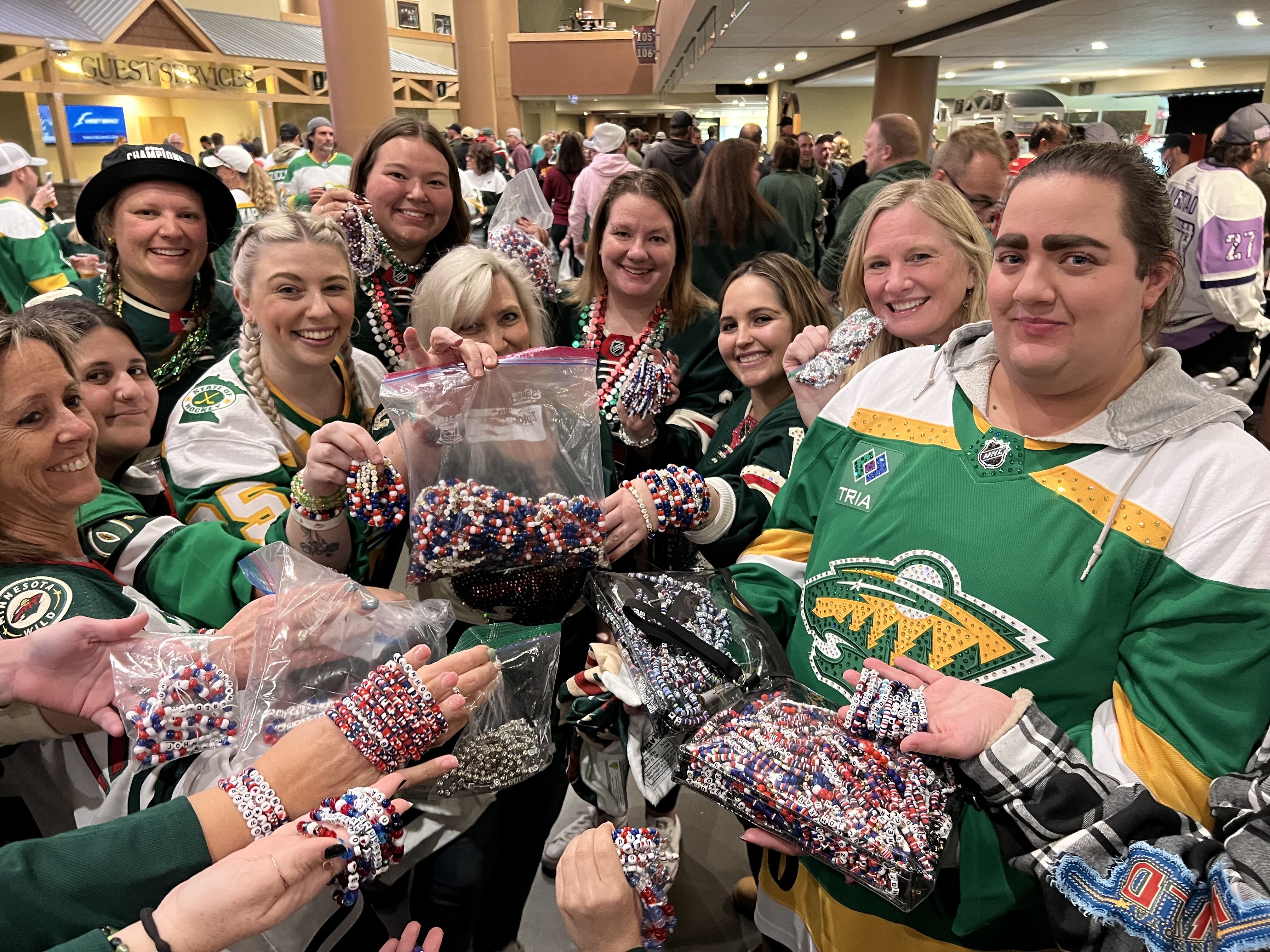 A group of women in green Minnesota Wild jerseys hold up plastic baggies full of red, white and blue beaded friendship bracelets