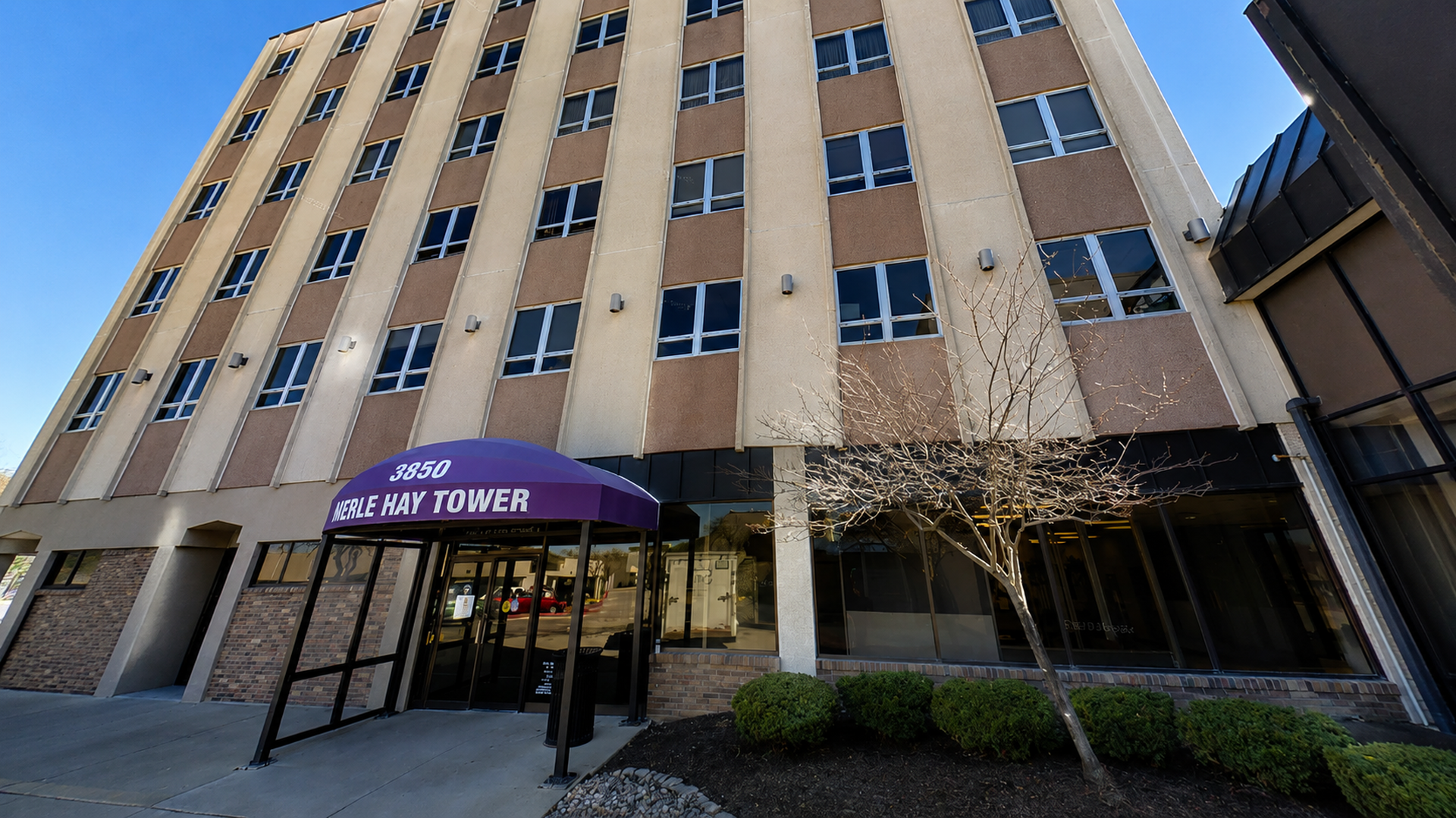 Wide view of a tall beige office building with brown panels. A curved purple canopy over the glass entrance reads "3850 MERLE HAY TOWER"; a leafless tree and shrubs line the sidewalk.