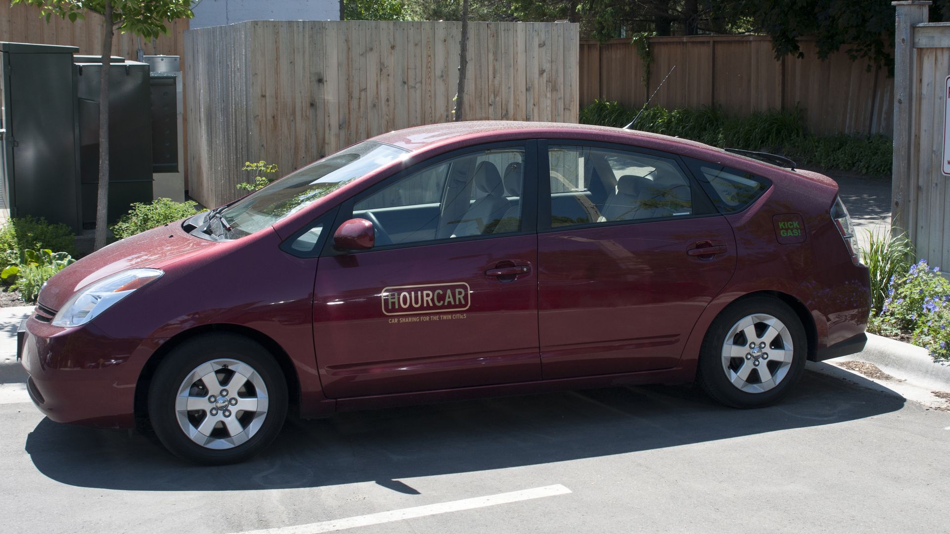 Maroon compact car parked along a paved lot with a wooden fence; two signs read "Hybrid Vehicle Parking Only" and "Reserved for Hourcar." Green trees and shrubs line the background.