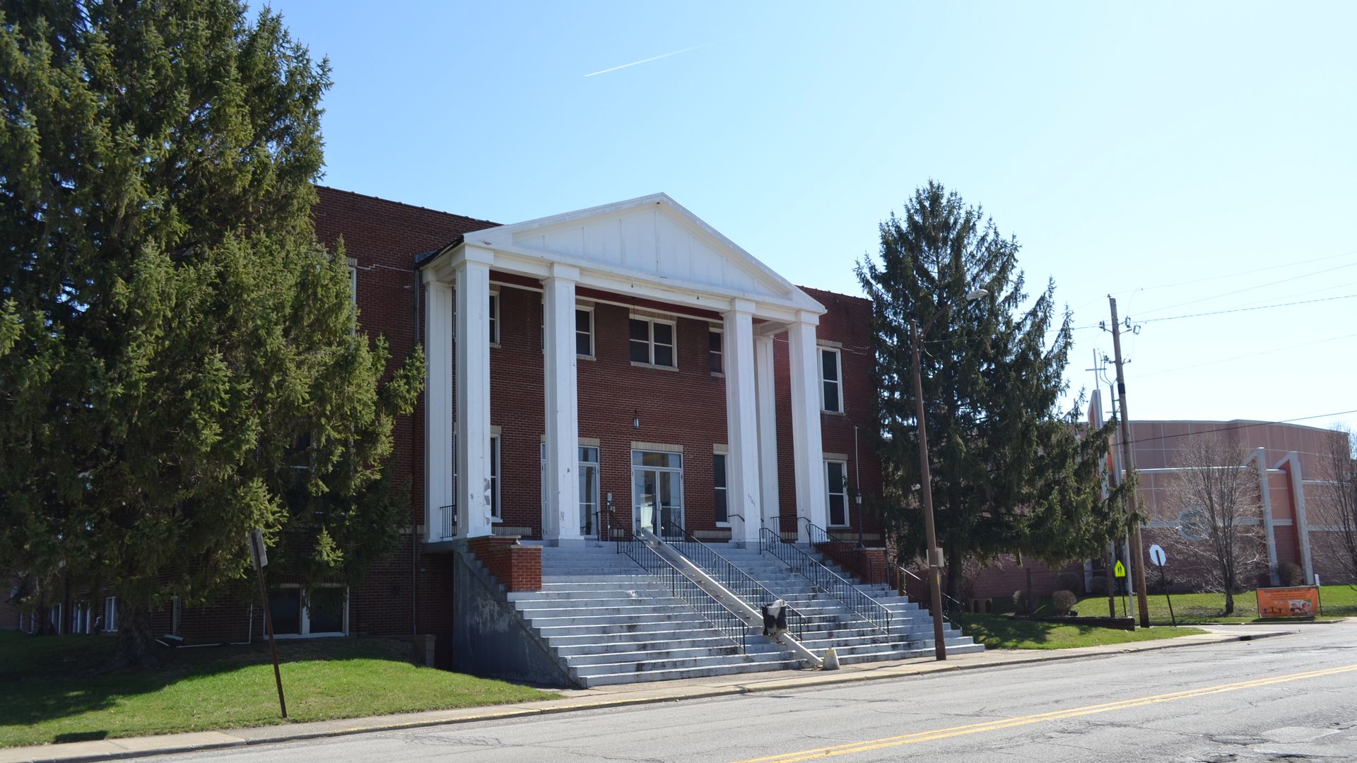 Red brick building with a white classical portico and tall columns, wide concrete steps with railings, flanked by large evergreen trees; a blue sky with a contrail above a quiet street.