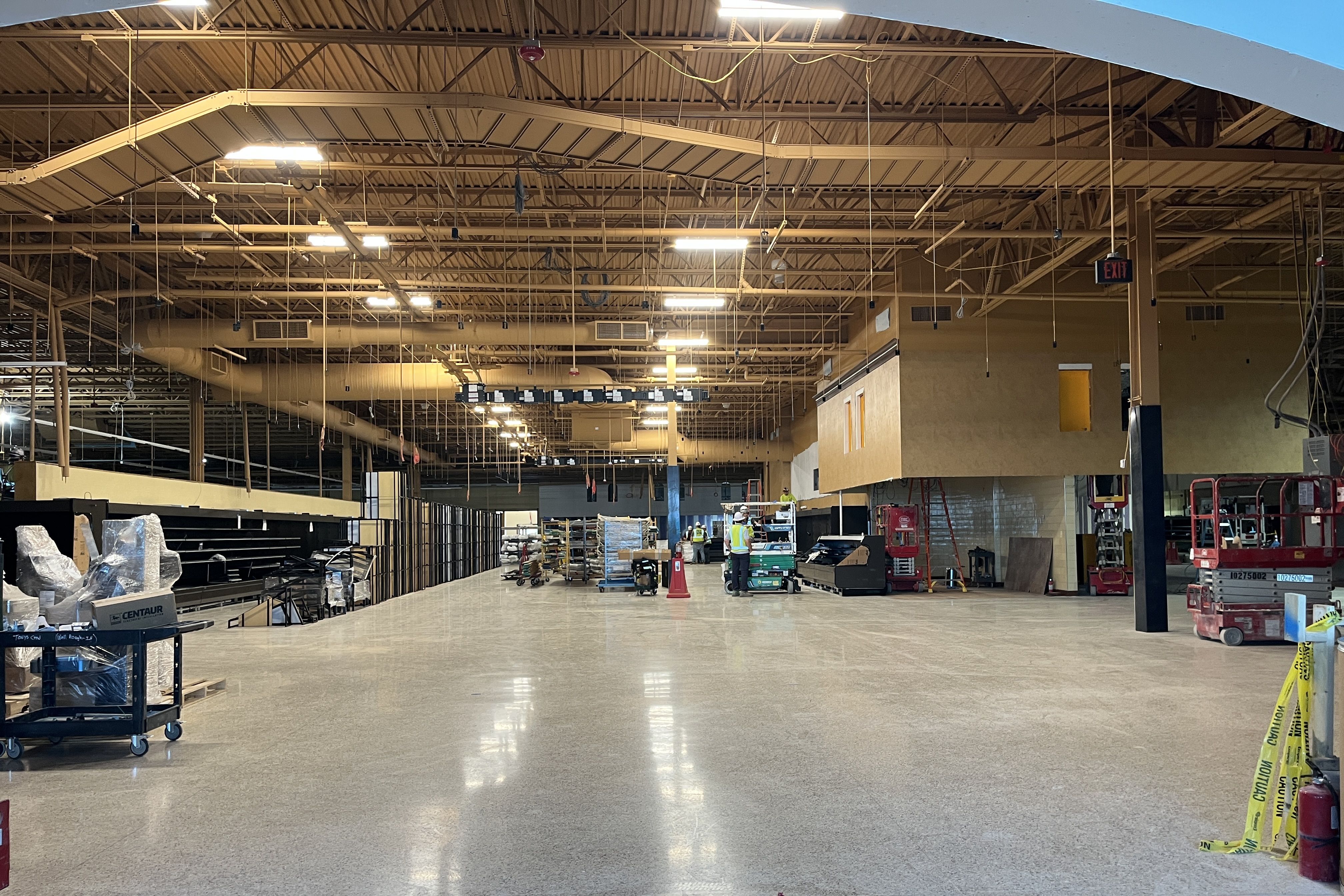 Interior of a large unfinished warehouse with exposed metal roof, hanging cords and lights, workers in safety vests near center, pallets and equipment, and yellow caution tape in the foreground.