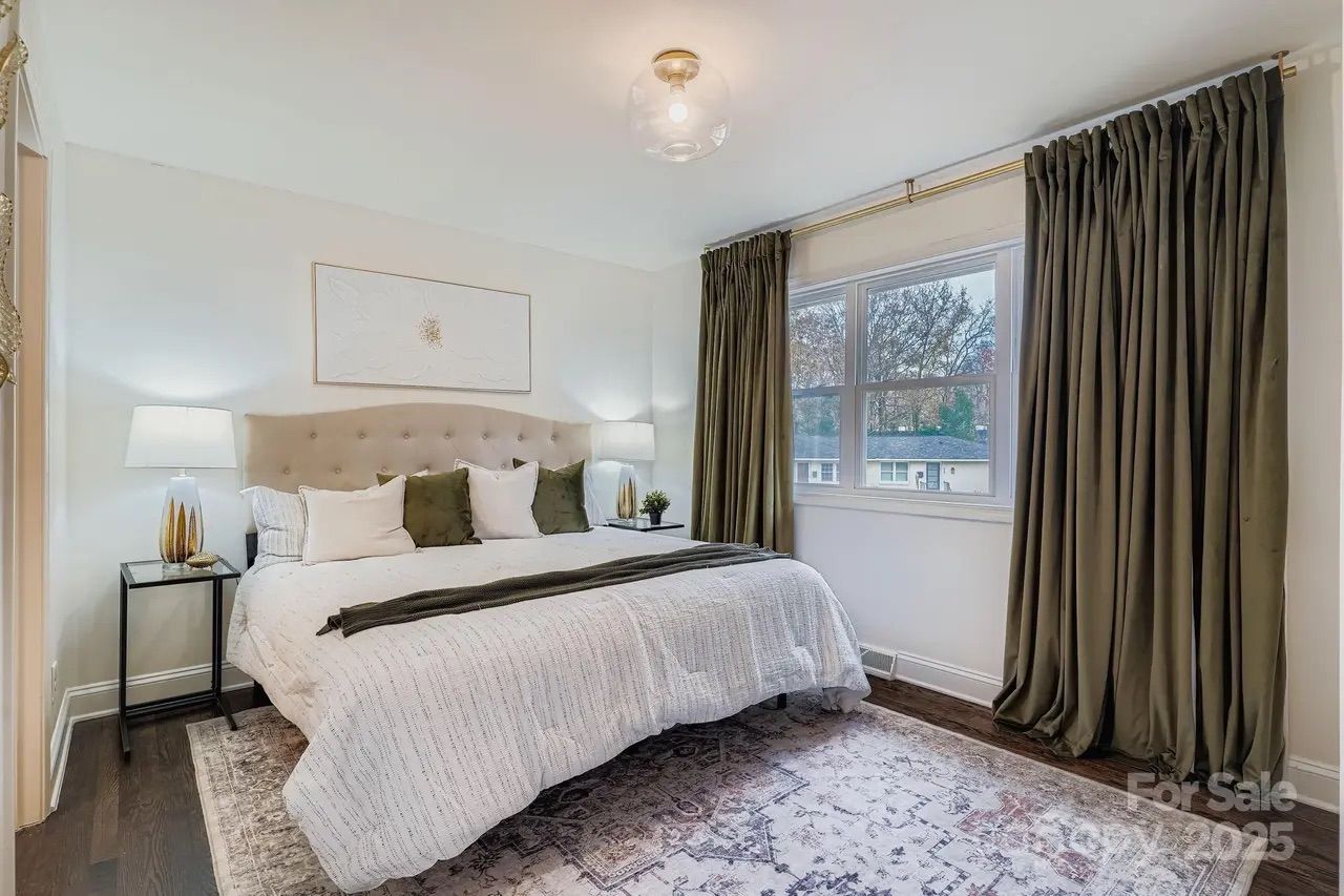 Bright bedroom with white walls, large bed with beige headboard, white and dark green pillows, white bedding, two lamps, dark green curtains, a patterned rug, and a window showing trees outside.