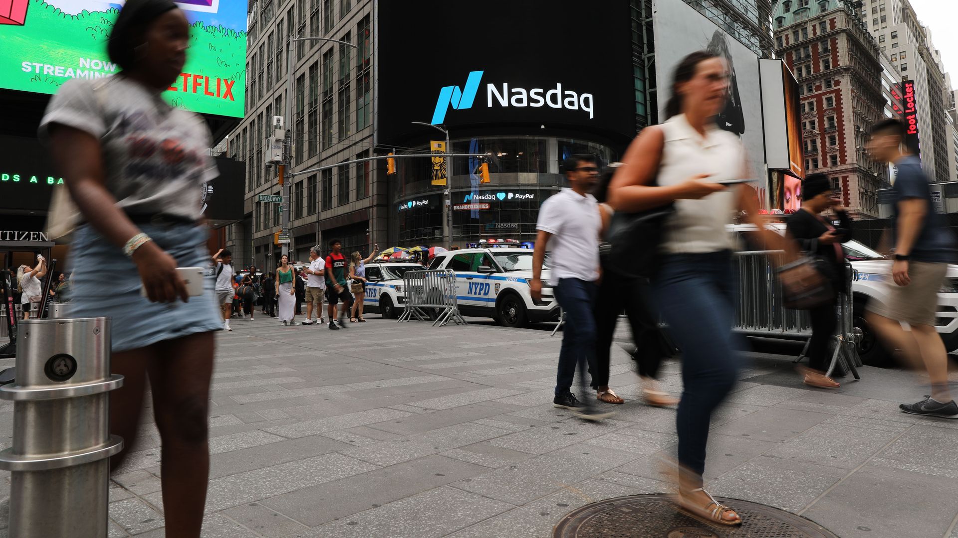 In this image, people rush by a digital NASDAQ sign in New York City.