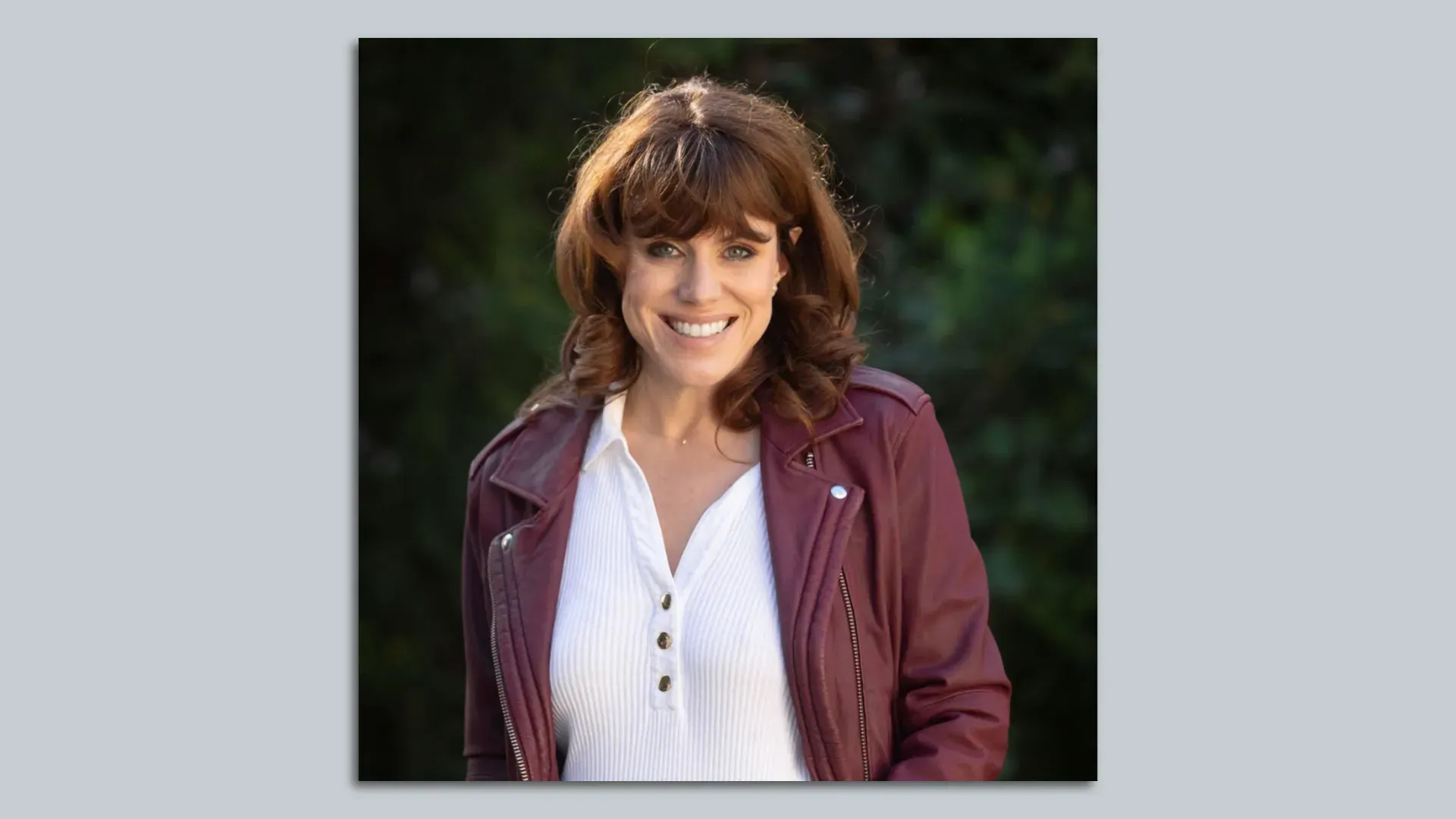 Smiling woman with shoulder-length brown hair, wearing a white button-up shirt and burgundy jacket, outdoors with a blurred green background.