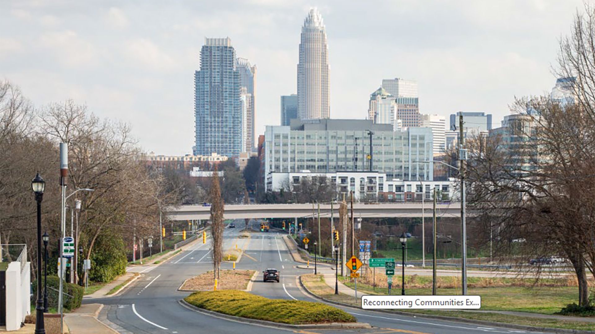 City skyline with tall glass towers behind a road and overpass; bare trees frame the foreground, while street lamps line the road and a green highway sign says Reconnecting Communities.
