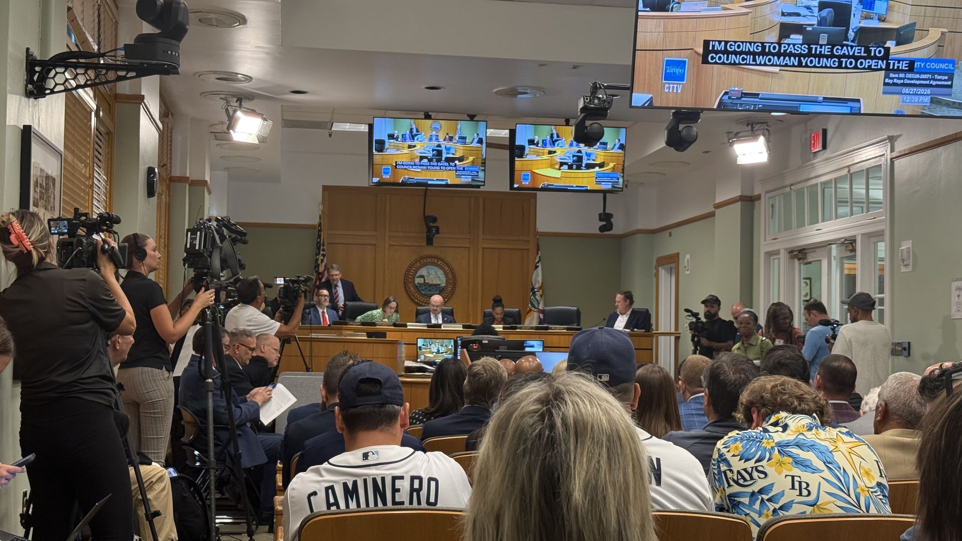 A crowded government chamber with reporters and cameras on the left, officials at a long desk, and overhead monitors showing the council meeting; audience, including people with Tampa Bay Rays jerseys and shirts, fill the foreground.