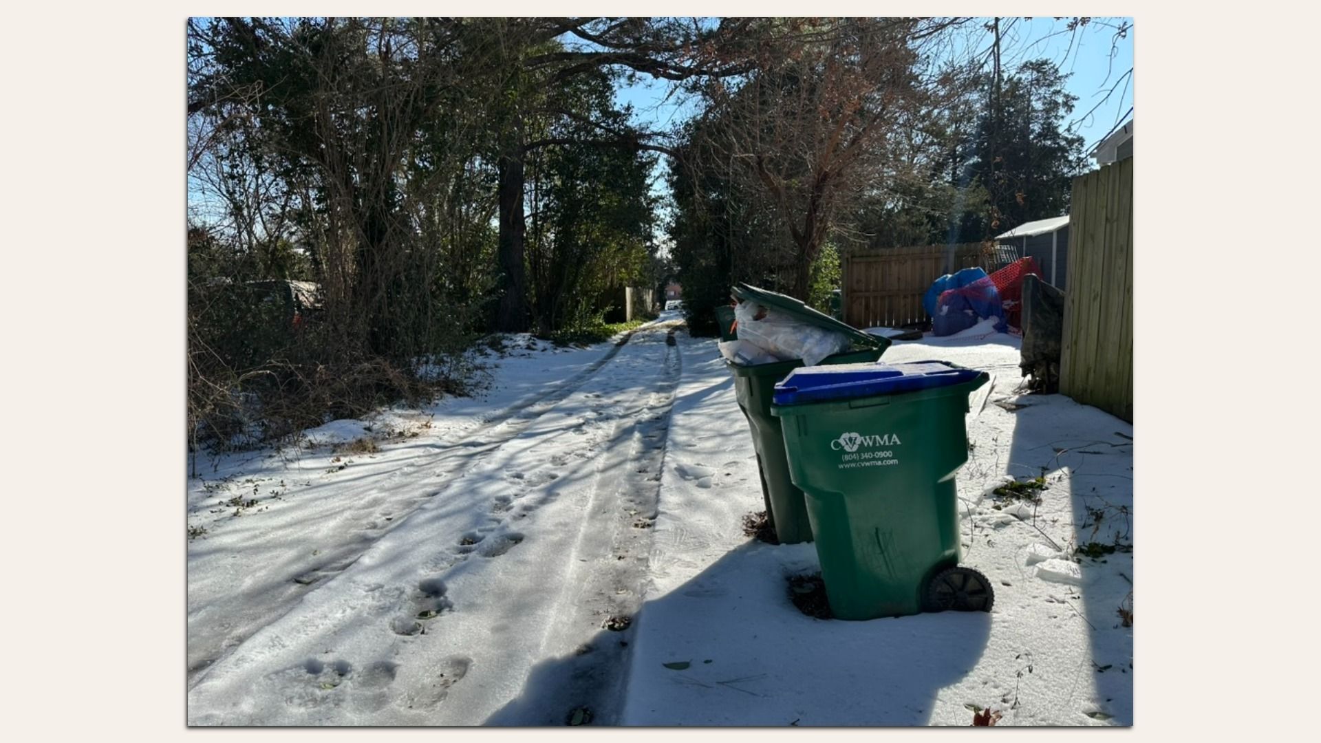 Snow-covered alley with tire and foot tracks, lined with bare trees on the left and a wooden fence on the right, with two green trash bins labeled "CVWMA" in foreground.