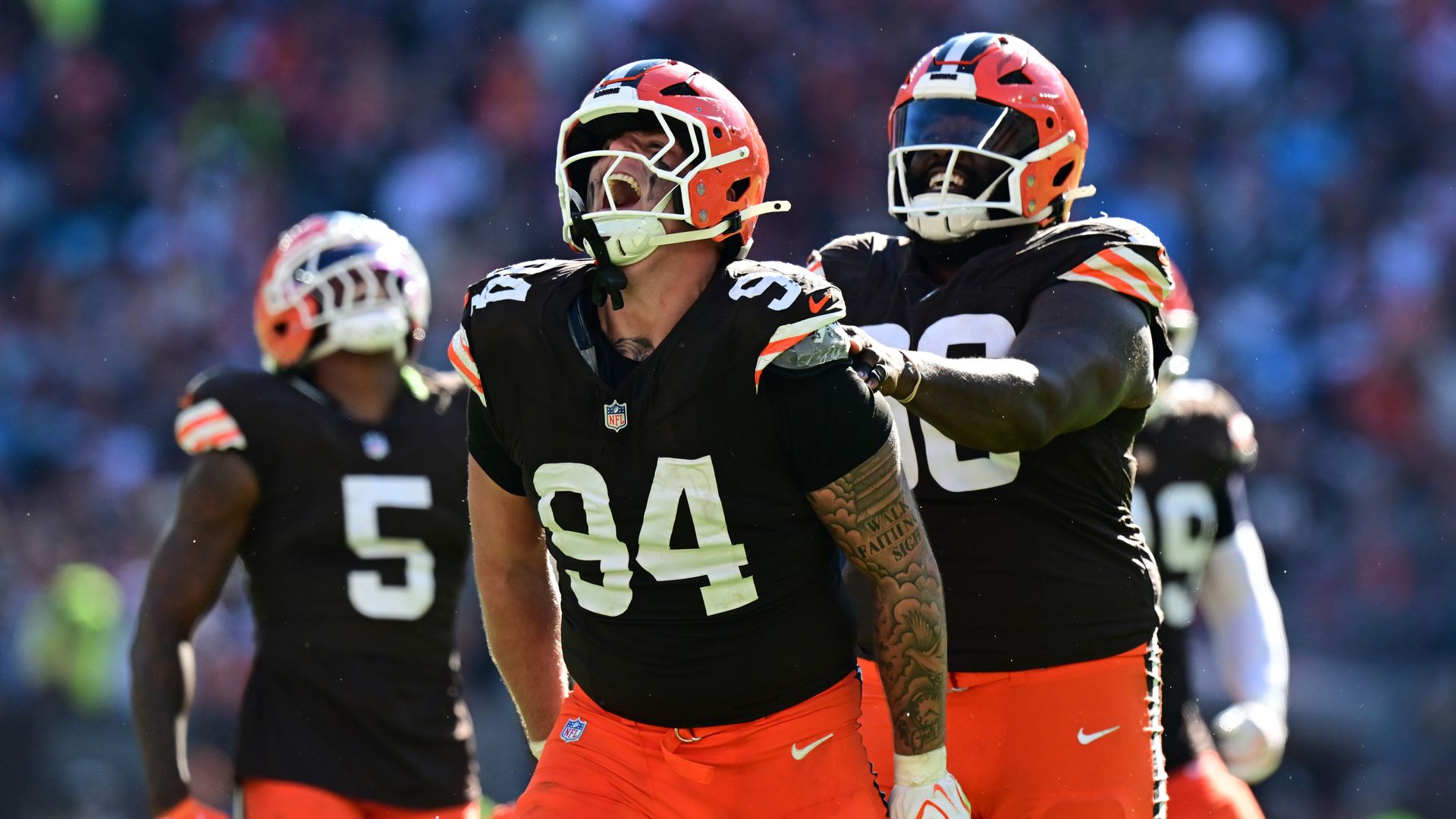 Two Cleveland Browns players in black jerseys and orange pants celebrate on the field, hugging each other under orange helmets, as a blurred stadium crowd watches.