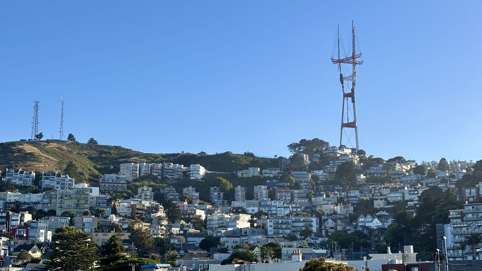 Blue sky over a hillside neighborhood with apartment buildings; a tall red-and-white telecom tower stands on the right, flanked by two smaller towers on the left.