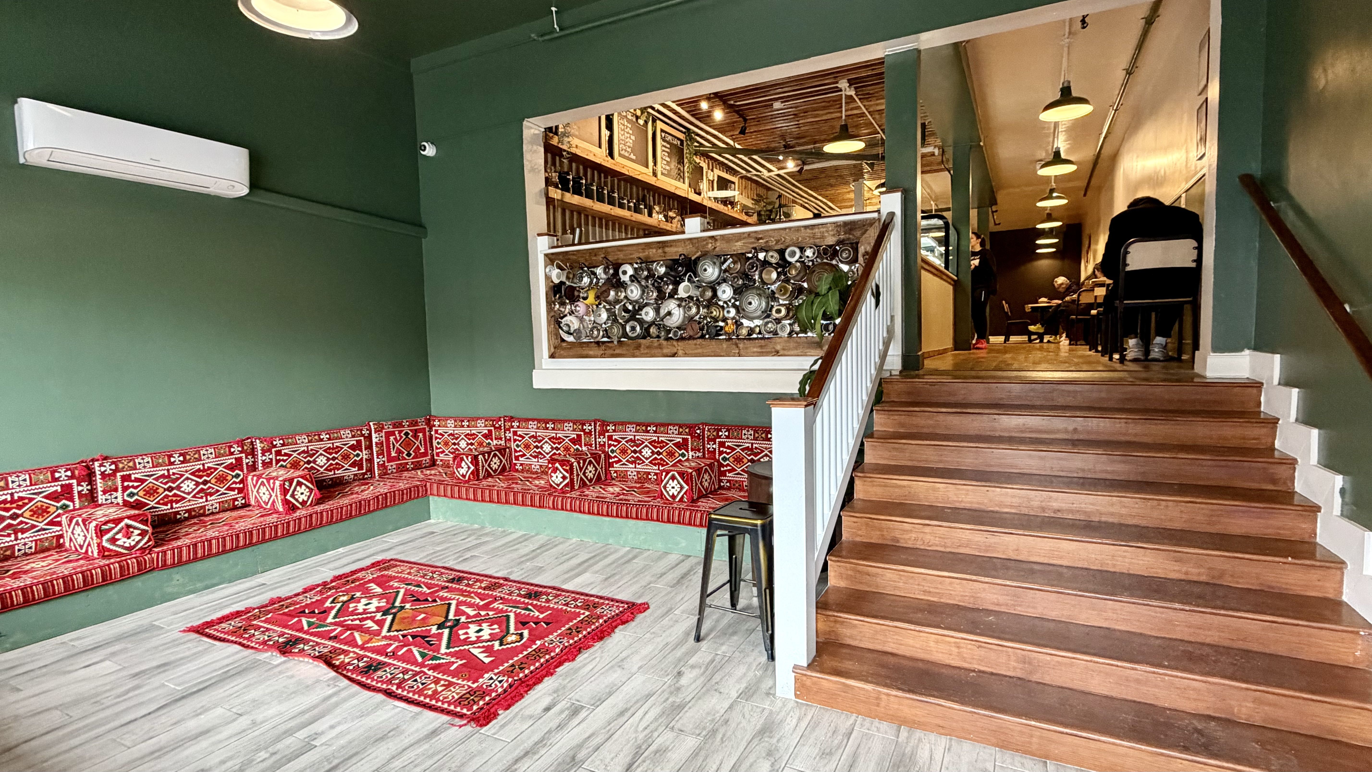 Indoor seating area with green walls, a red patterned sofa and rug, wooden stairs leading to a cozy café space with warm lighting and people seated inside.