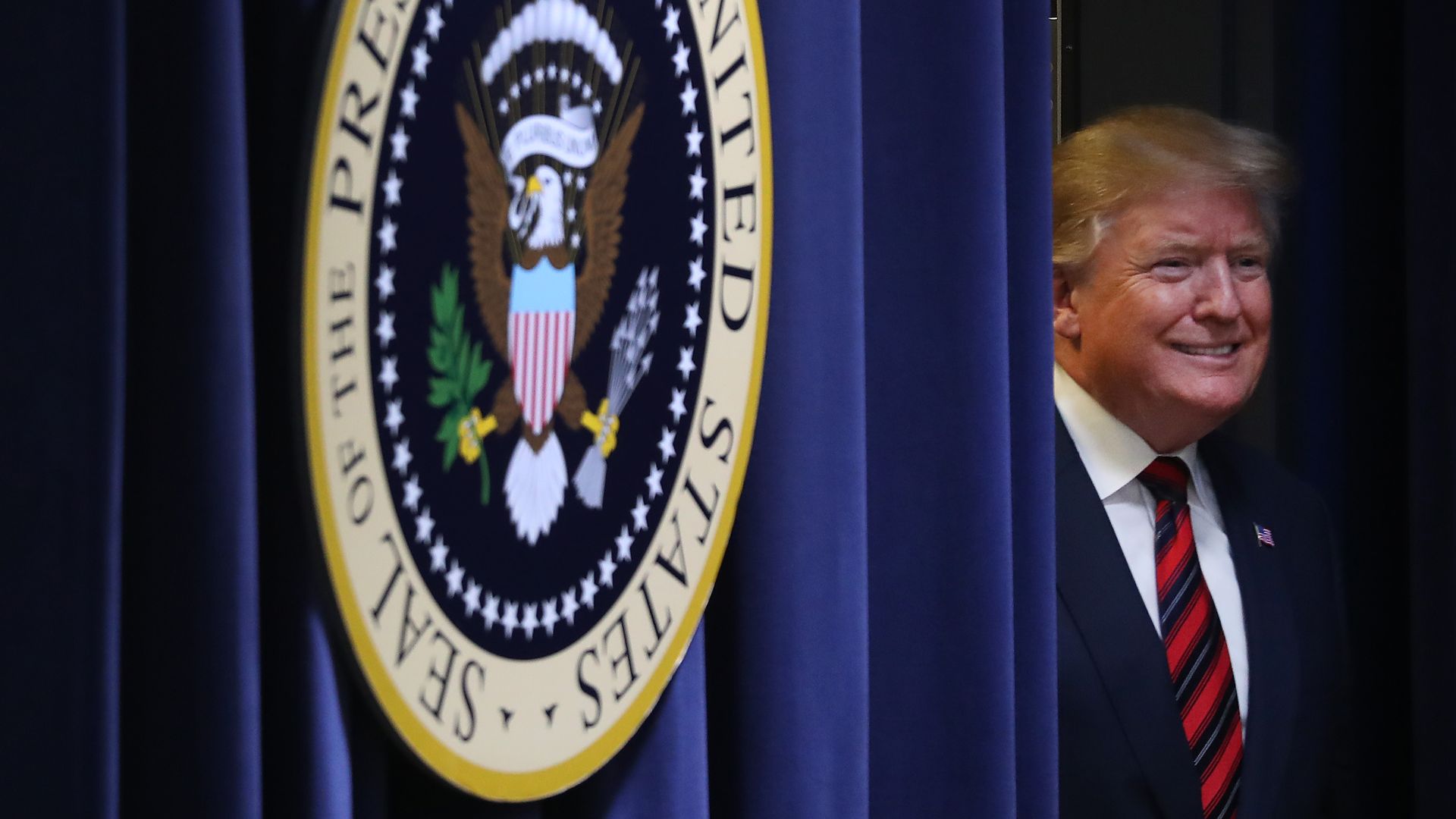 In this image, Trump smiles next to a heavy blue curtain next to the presidential seal. 