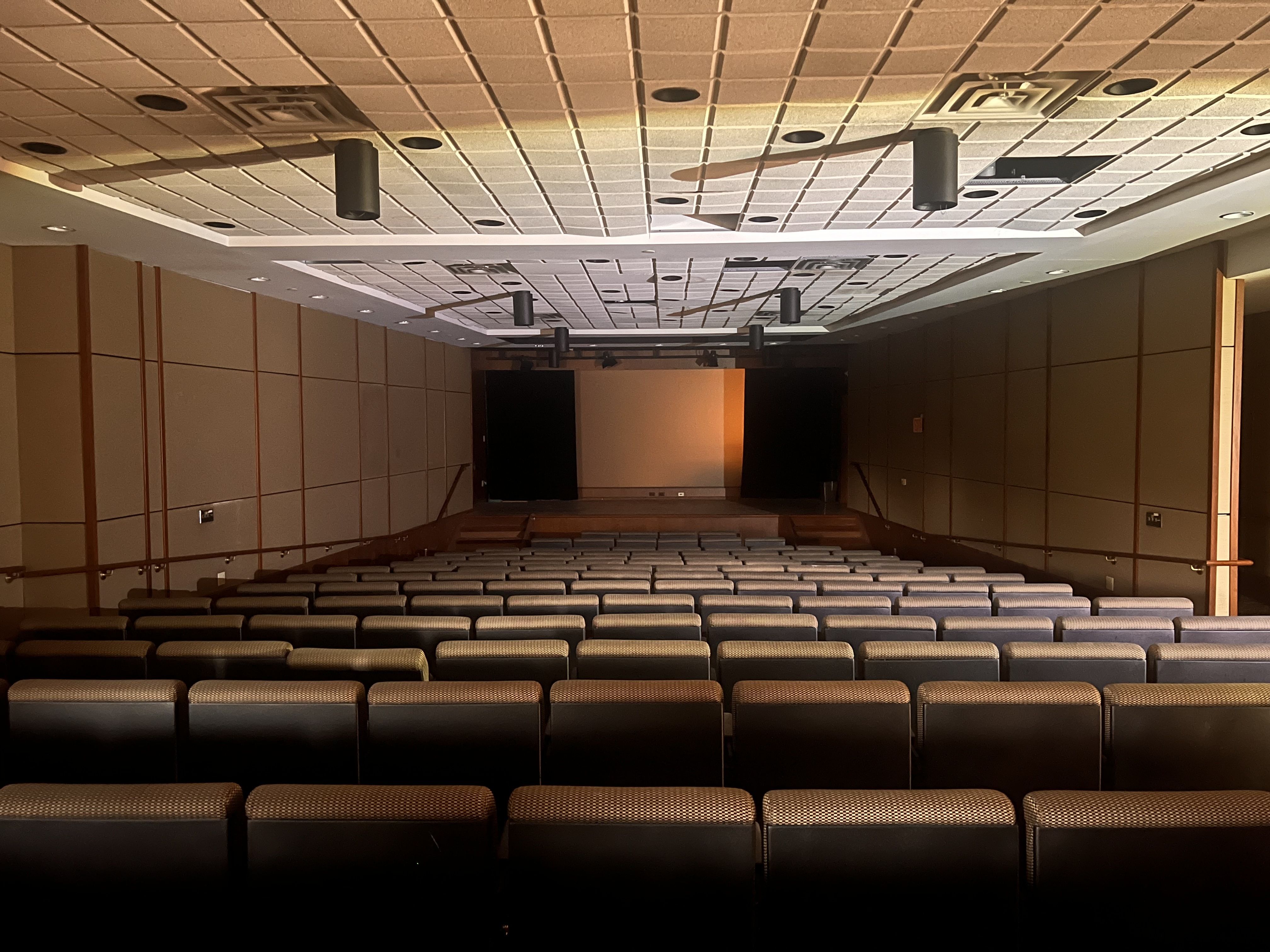 Empty auditorium with rows of gray upholstered seats facing a distant stage. Brown paneled walls, a grid ceiling with vents and hanging black speakers, warm stage lighting.