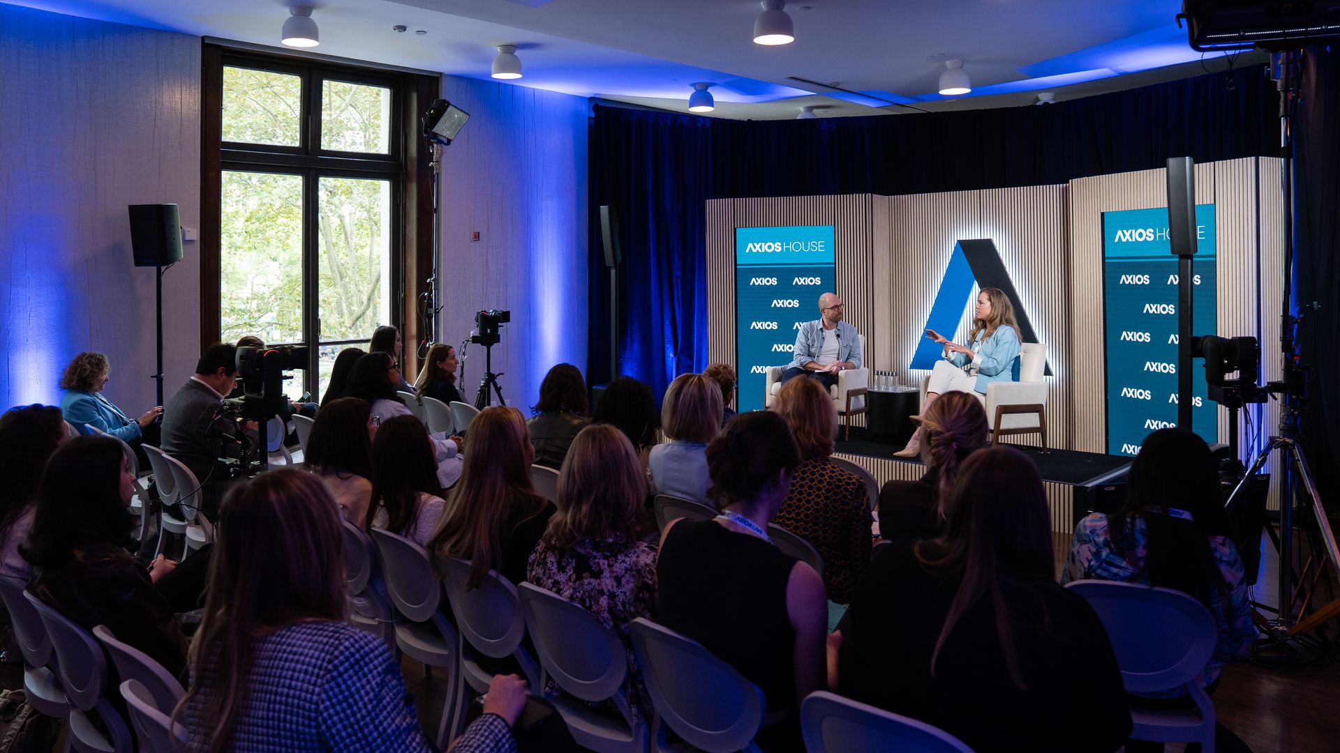 Panel discussion on a stage at Axios House: two speakers sit in chairs under blue lighting, tall backdrops with Axios House logos, an audience watches as cameras record, a large window left.