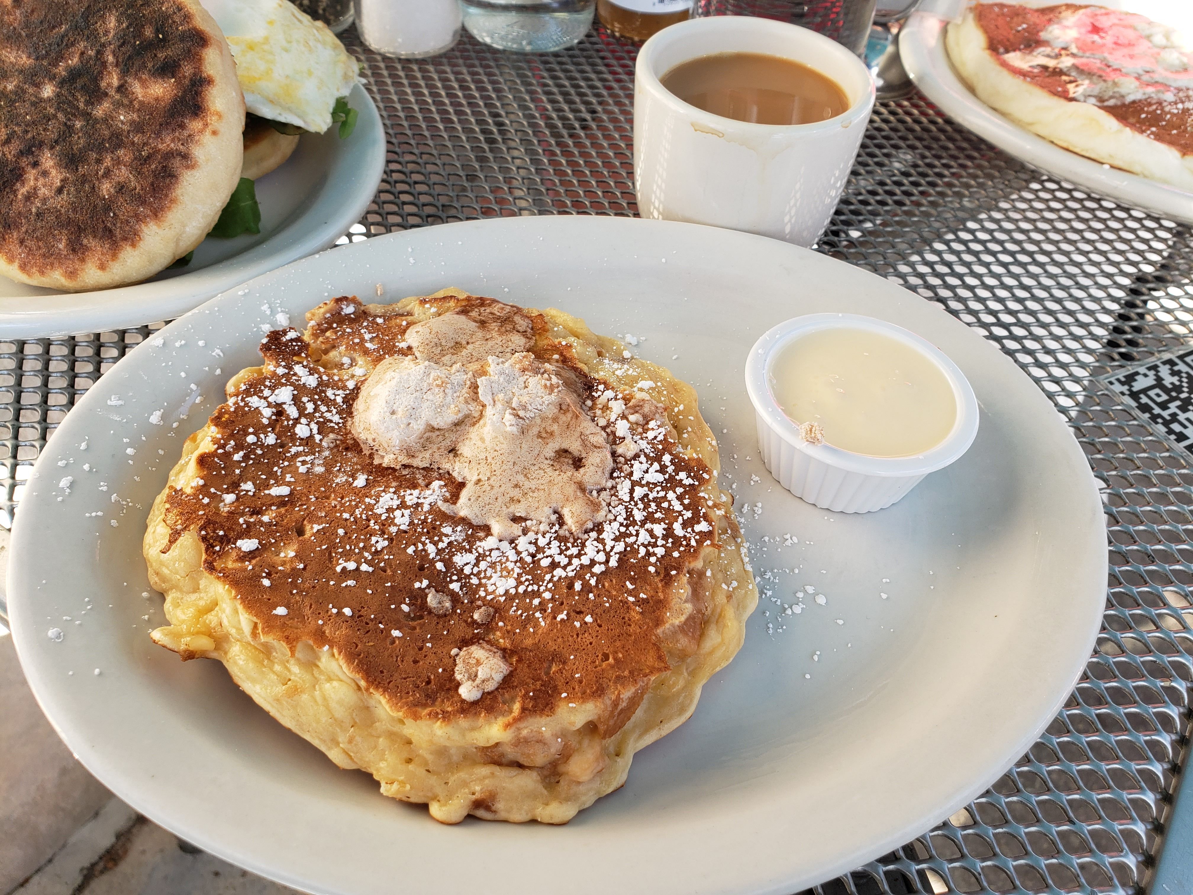 Outdoor meal on a metal table: a plate with two thick pancakes dusted with powdered sugar in the foreground, a white mug of coffee nearby, another plate with a bagel sandwich and a syrup cup.