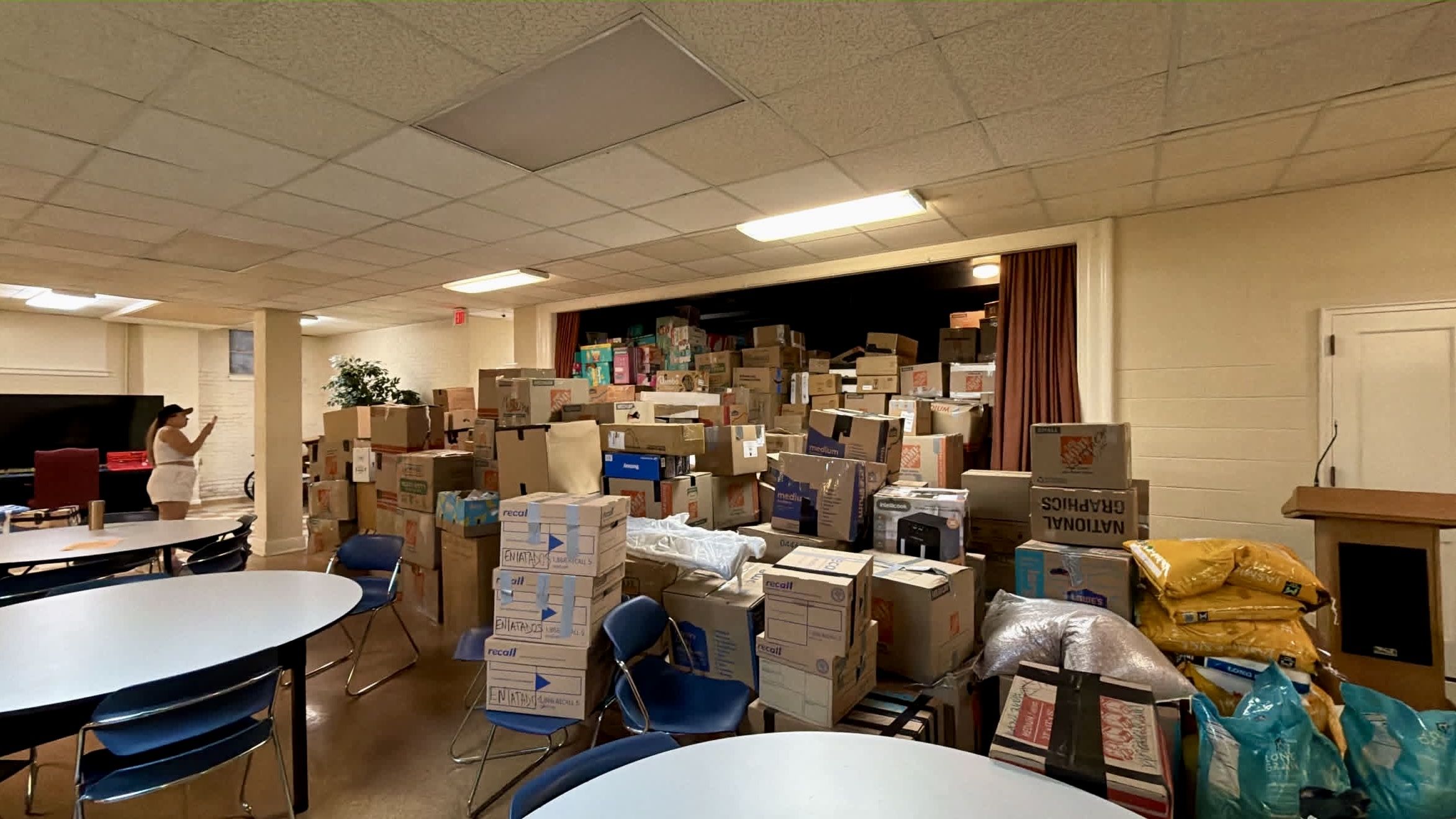 Room filled with stacked cardboard boxes and supplies; a person in white with a cap stands left, taking a photo. Round tables and blue chairs sit in the foreground under fluorescent ceiling lights.