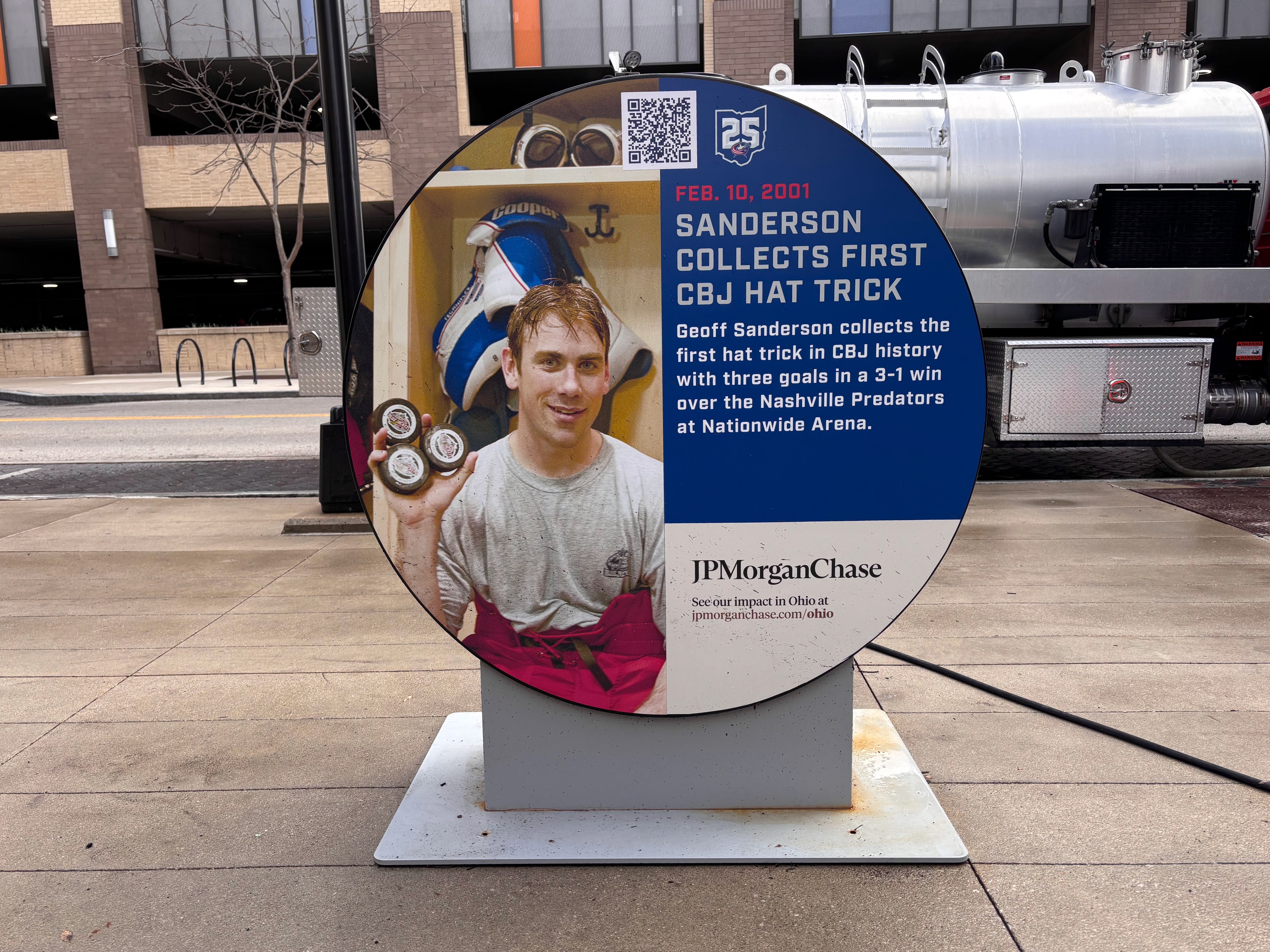 Round informational display with a photo of a man holding three hockey pucks on the left and blue text about the history of Geoff Sanderson scoring the first CBJ hat trick on the right.