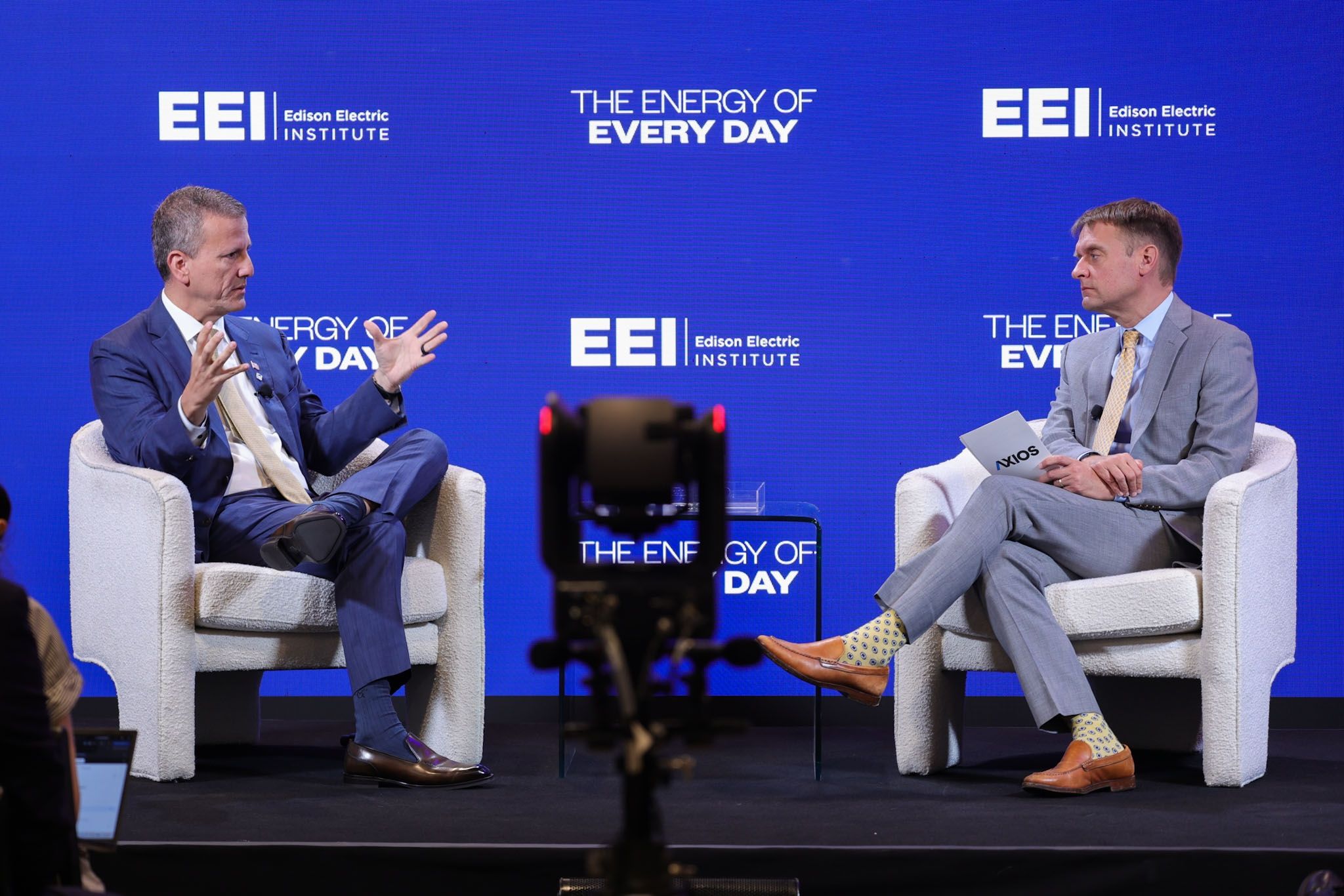 Two men in suits sit in white chairs on a blue stage with EEI Edison Electric Institute logos. The backdrop reads "THE ENERGY OF EVERY DAY." A camera frames the interview in the foreground.