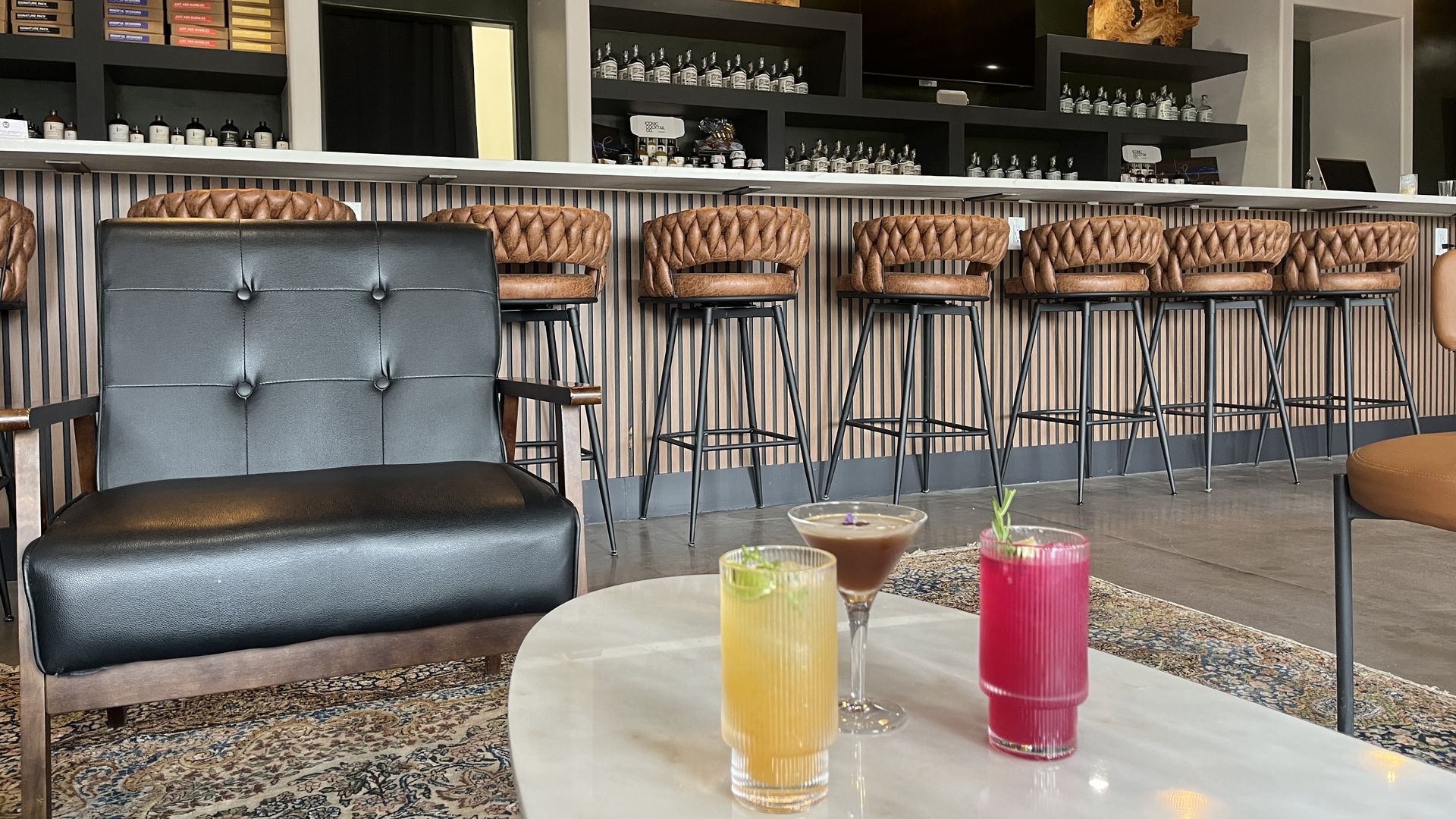 Interior of a modern bar with black leather chair, brown leather bar stools along the counter, hanging pendant lights, and three colorful cocktails on a white marble table.