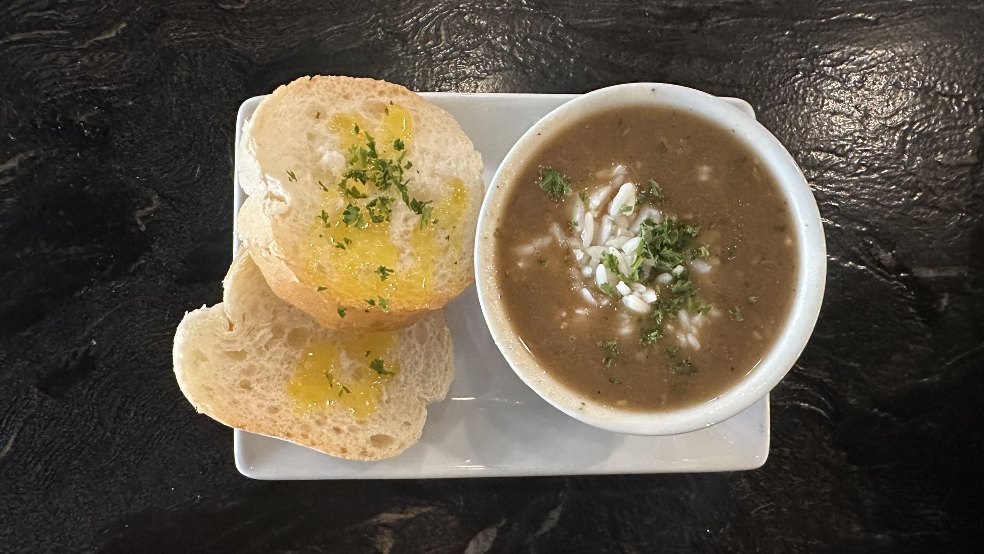 A cup of gumbo sits on a dark stone countertop on top of a rectangle plate. The plate also has two bread rounds.