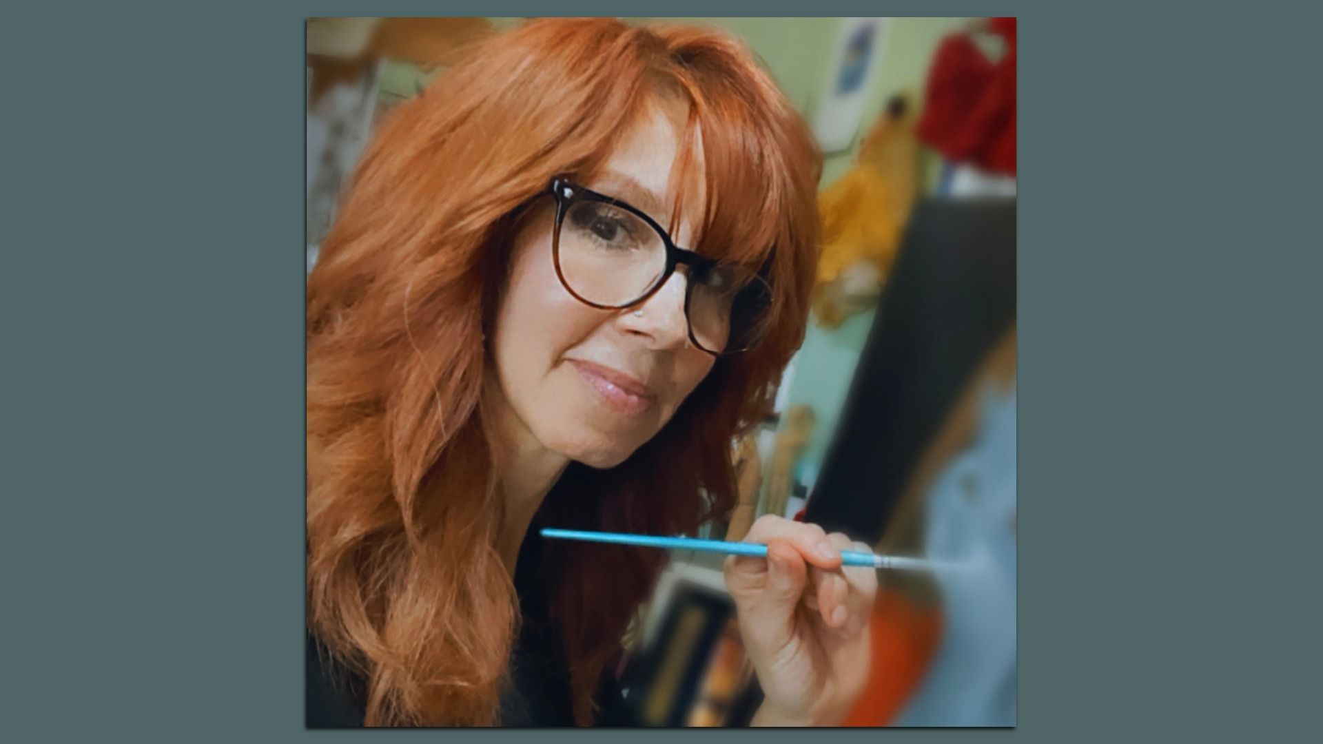 Red-haired woman with glasses holding a paintbrush, smiling gently, with an art studio background blurred behind her.