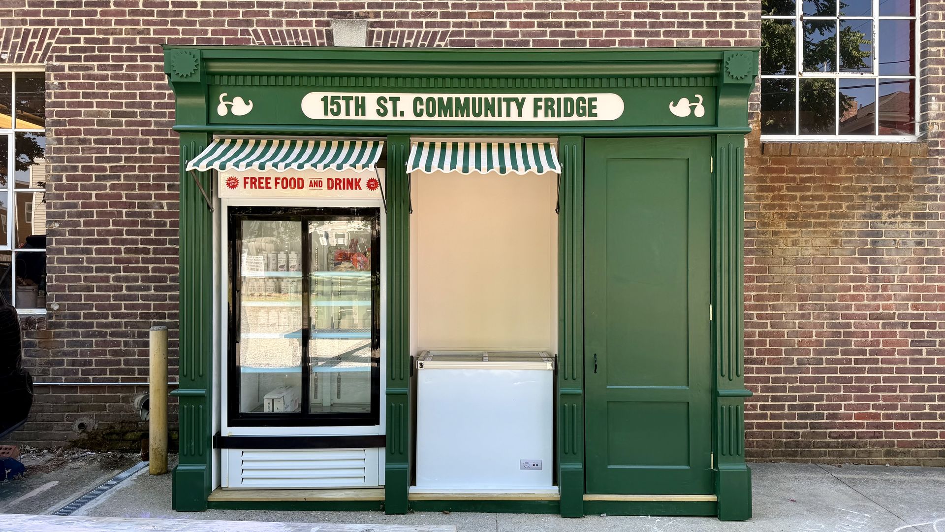 Green storefront on a brick building, sign reads "15th St. Community Fridge." Striped green-and-white awnings shade a glass fridge; a red banner proclaims "FREE FOOD AND DRINK."